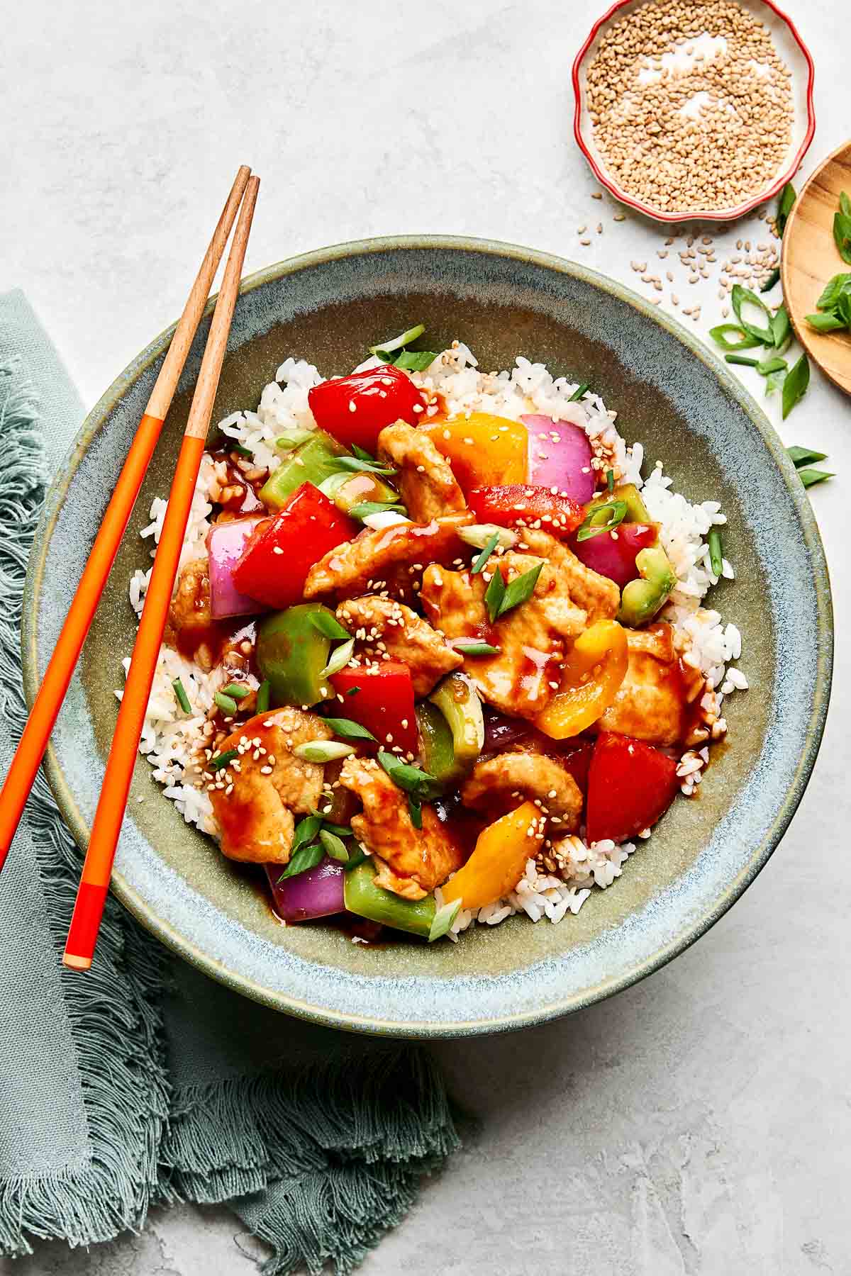 A bowl of white rice topped with sweet and sour chicken, colorful bell peppers, red onion, and sesame seeds, with chopsticks resting on the bowl and garnishes nearby.