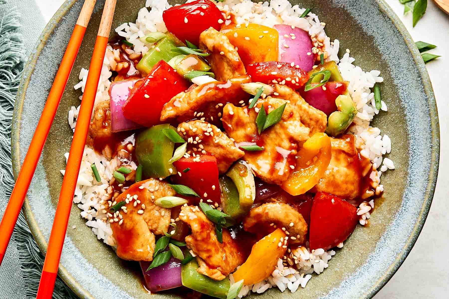 A bowl of white rice topped with sweet and sour chicken, colorful bell peppers, red onions, and sauce, garnished with sesame seeds and chopped green onions, with red chopsticks resting on the bowl.