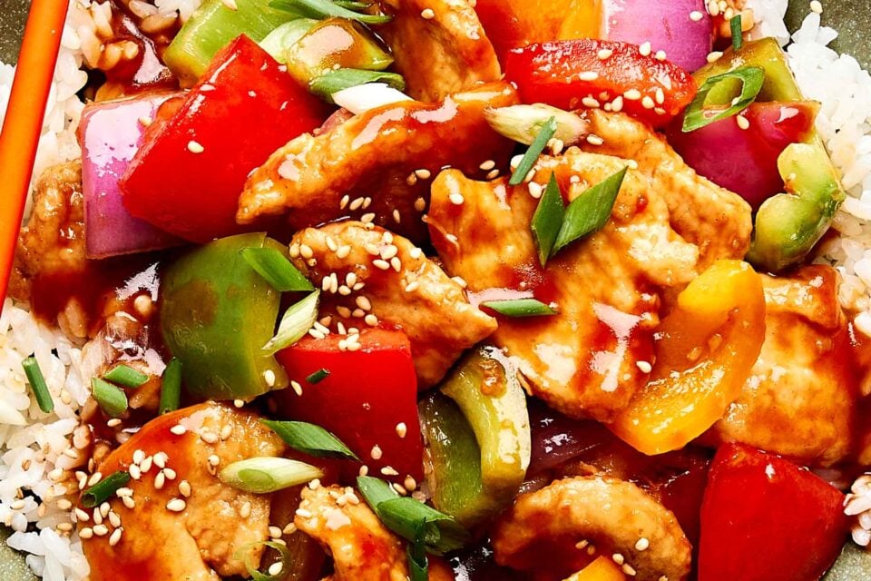 A close-up of a colorful sweet and sour chicken stir-fry with pieces of chicken, red and green bell peppers, onions, and scallions over white rice, topped with sesame seeds and a glossy sauce.