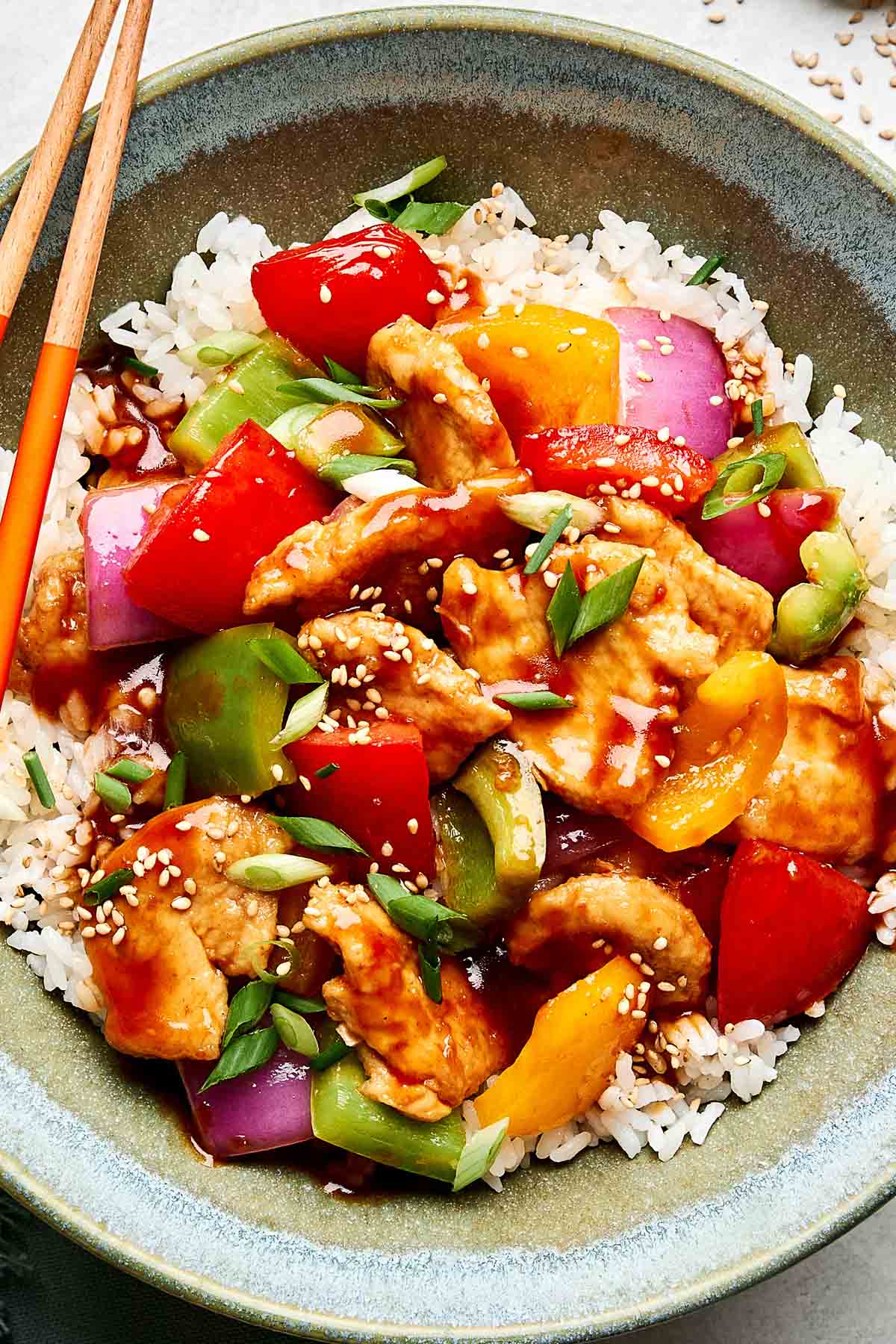 A bowl of white rice topped with sweet and sour stir-fried chicken, red and green bell peppers, purple onions, and a glossy sauce, garnished with sliced green onions and sesame seeds. Chopsticks rest on the side of the bowl.