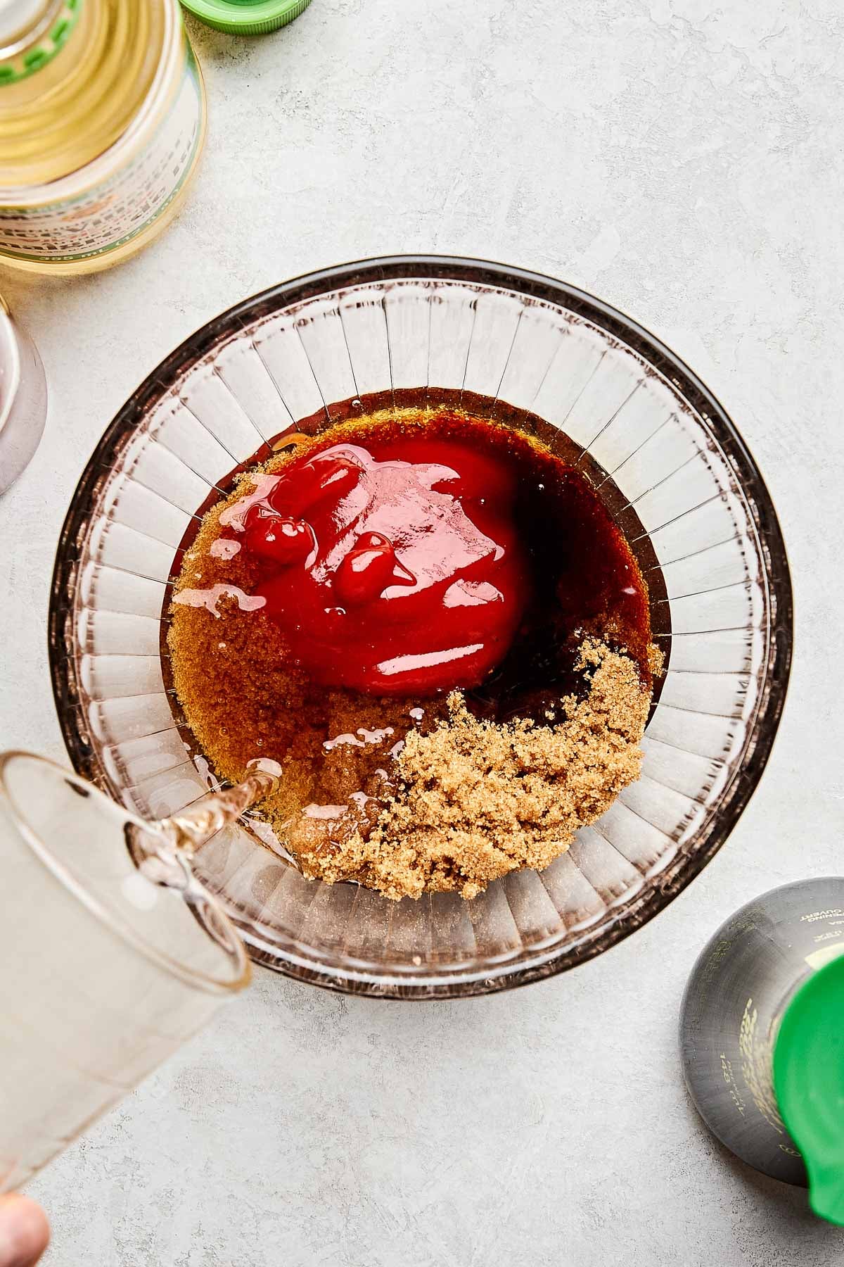 A glass bowl containing brown sugar, ketchup, and soy sauce, with a hand pouring a clear liquid from a small glass into the mixture, surrounded by various bottles on a light-colored surface.
