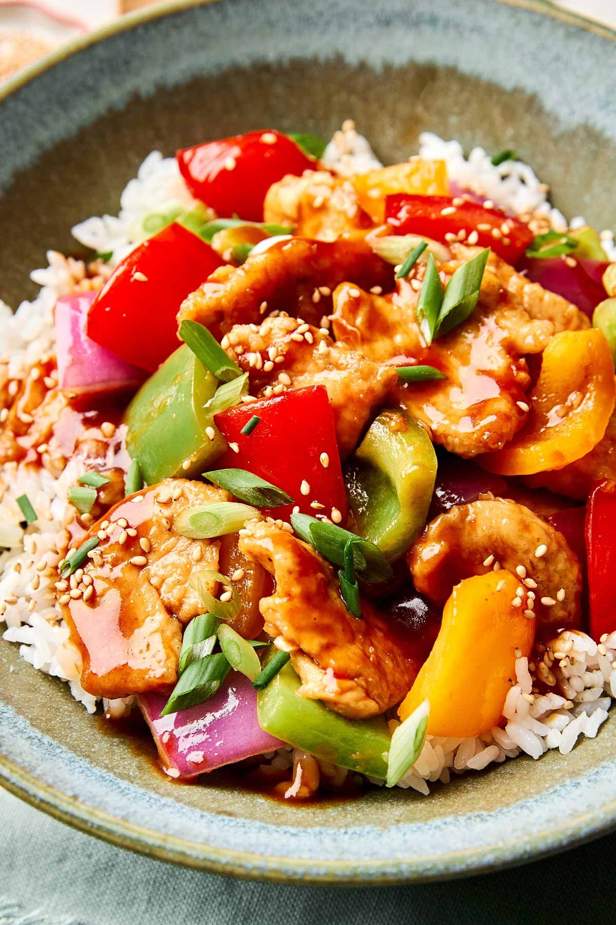 A bowl of white rice topped with sweet and sour stir-fried chicken, red and yellow bell peppers, green bell peppers, red onion, and chopped green onions, all coated in a glossy sauce and sprinkled with sesame seeds.