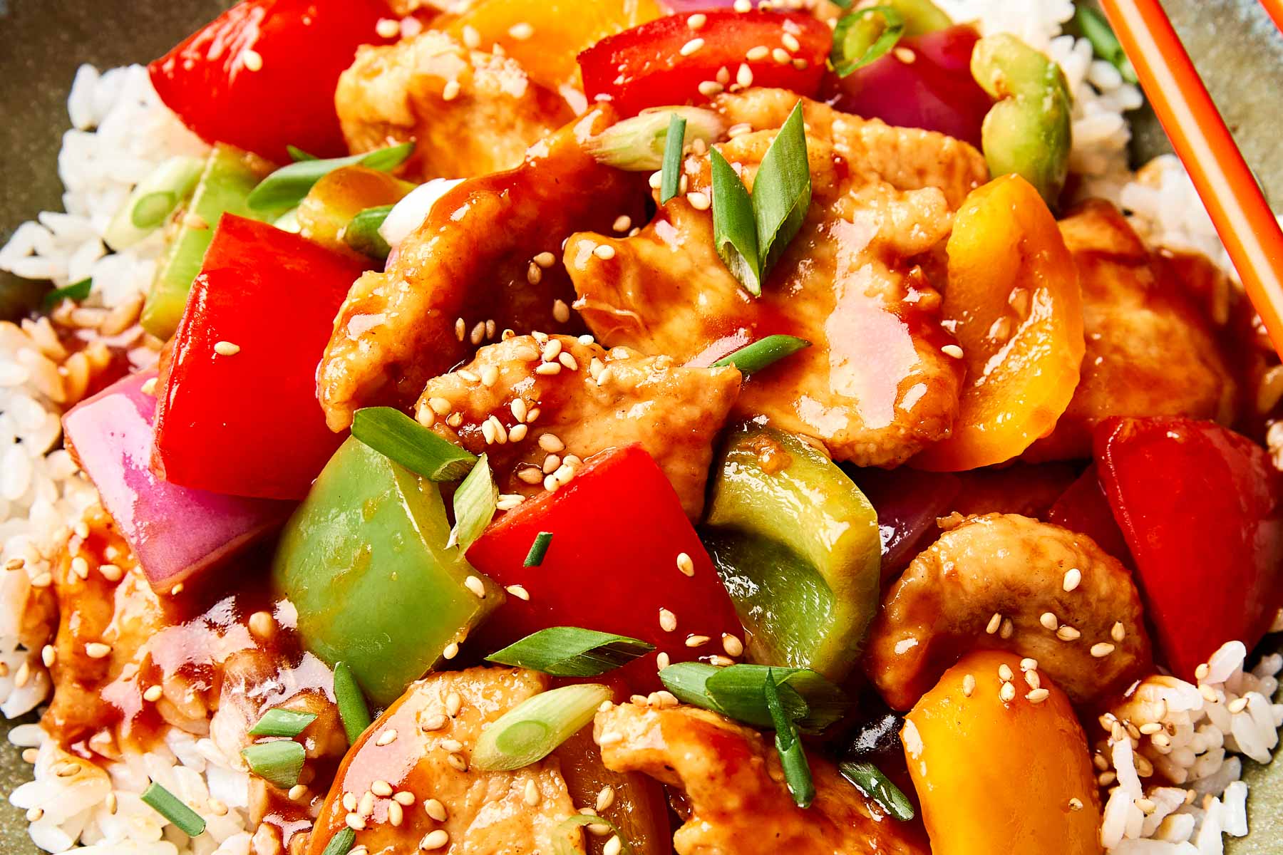 A close-up of sweet and sour chicken with bell peppers and onions, served over white rice, garnished with sesame seeds and sliced green onions.