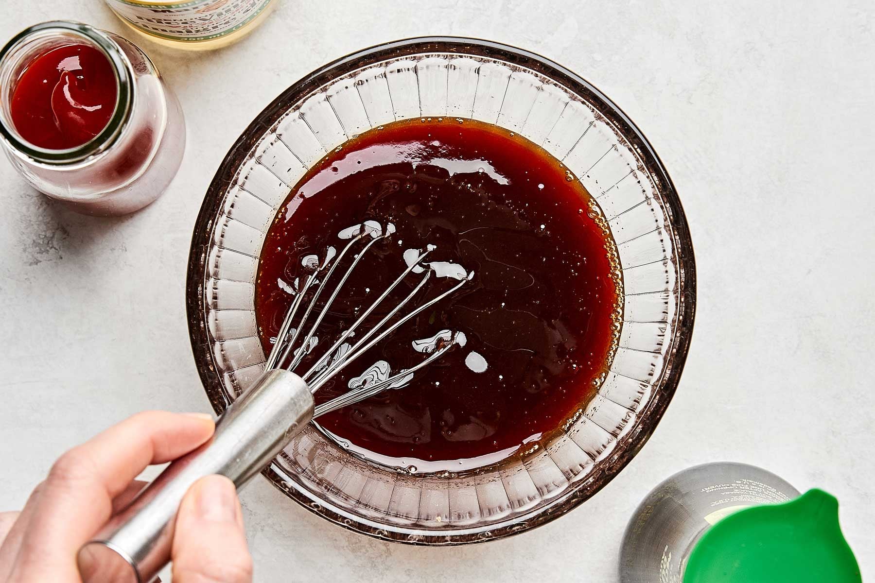 A hand uses a metal whisk to mix a dark brown sauce in a glass bowl. Surrounding the bowl are bottles containing red and yellow liquids, and a green-capped container. The setting is a light-colored countertop.