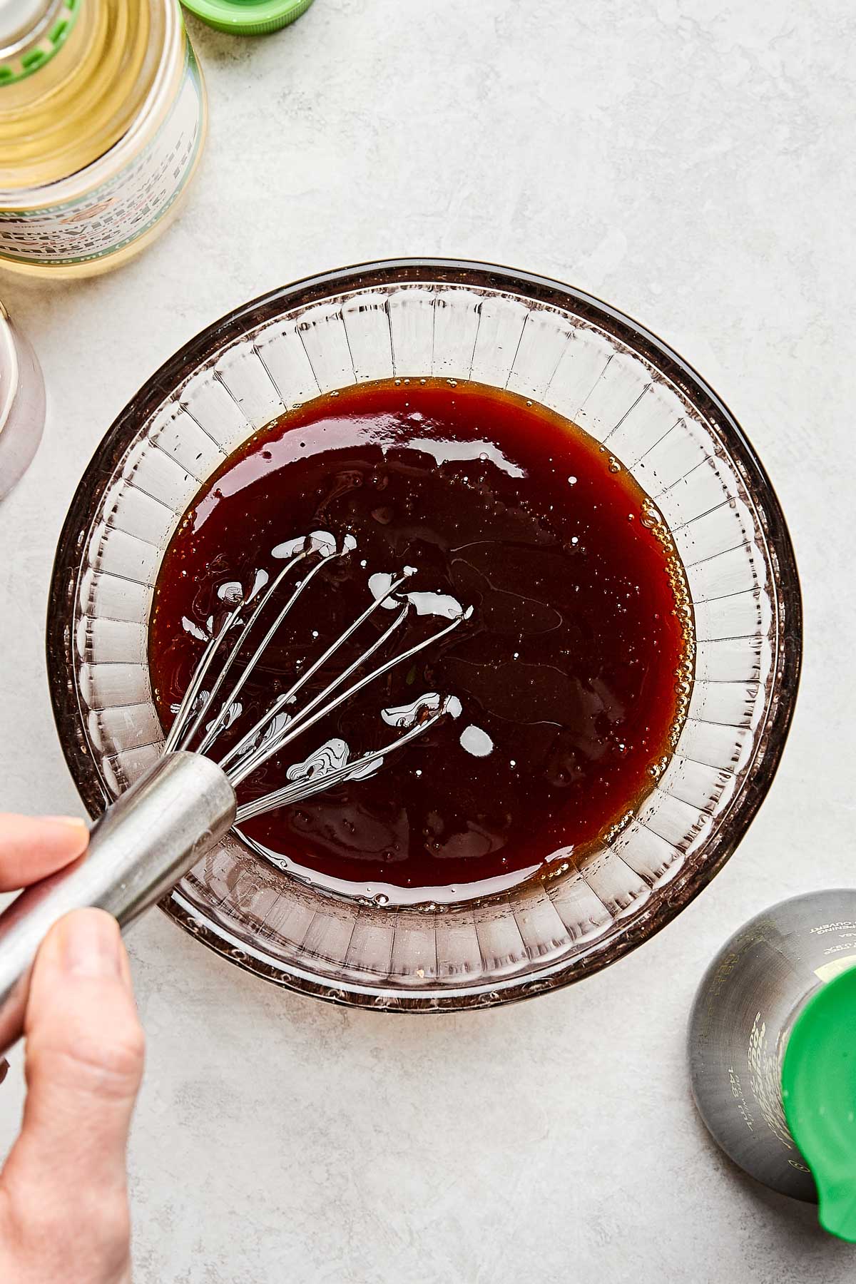 A hand holding a whisk is mixing sweet and sour sauce in a clear glass bowl on a light countertop, with bottles partially visible around the bowl.