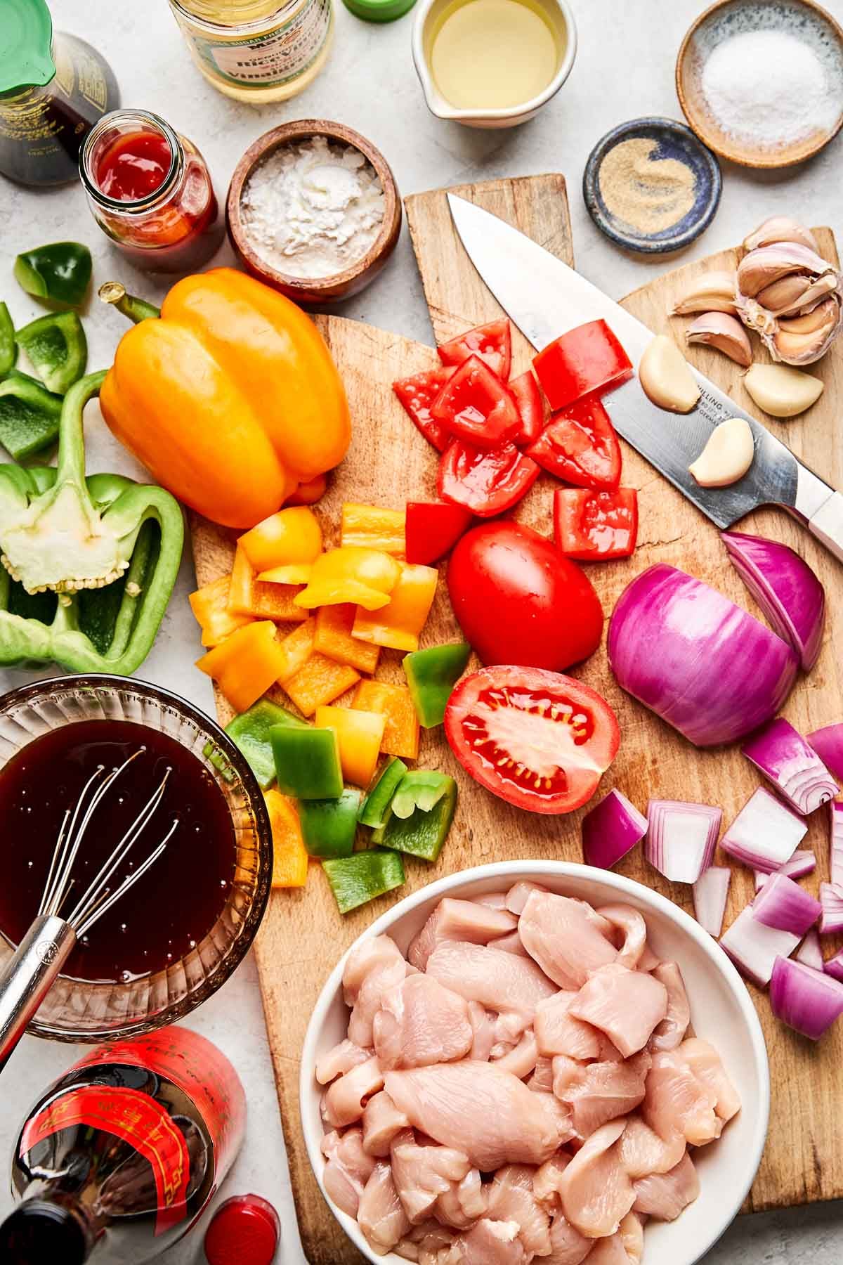 Chopped raw chicken, diced tomatoes, red onion, green and yellow bell peppers, garlic cloves, and various sauces and seasonings on a cutting board and table, ready for cooking.