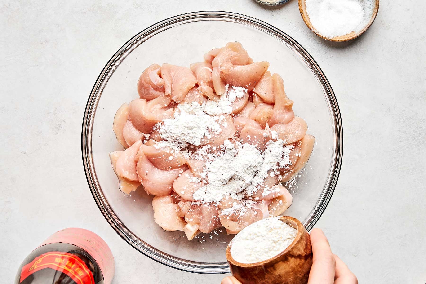 A hand sprinkles white powder, likely cornstarch, over raw chicken pieces in a glass bowl. A small bowl of salt and a bottle with a red cap are nearby on a light surface.