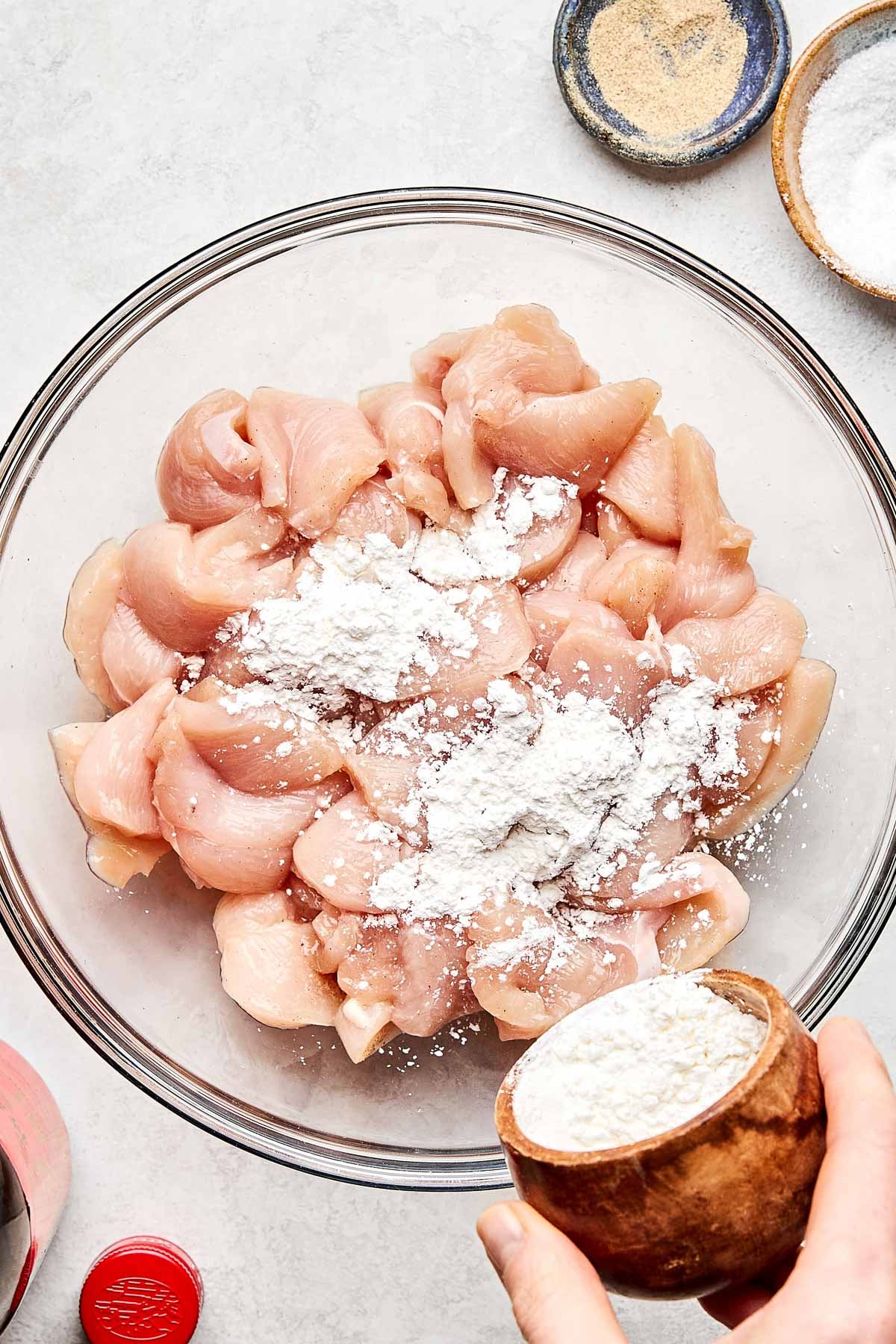 A hand sprinkles flour onto raw chicken pieces in a glass bowl. Nearby, small bowls hold various seasonings, and a red container is partially visible on a light-colored surface.