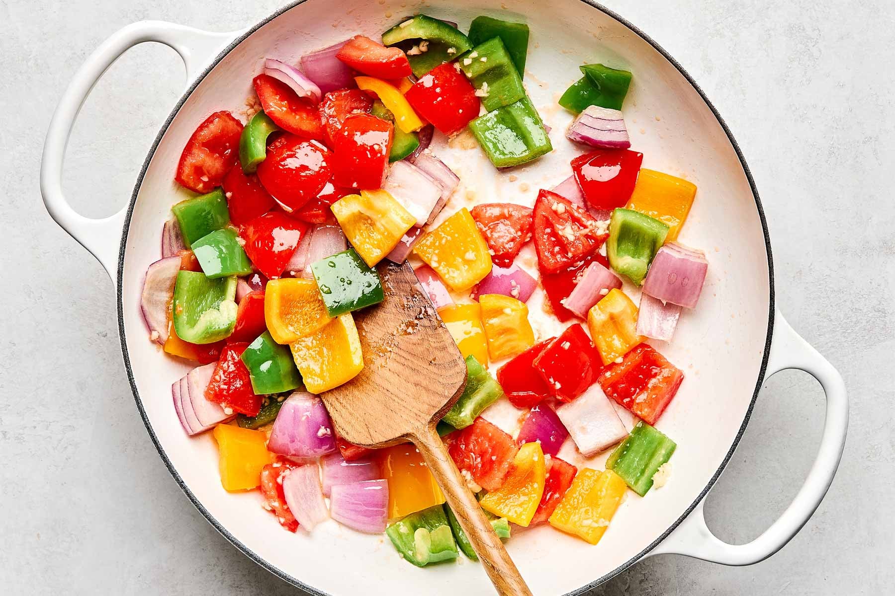 A white skillet contains chopped red, yellow, and green bell peppers, red onions, and tomatoes being sautéed. A wooden spatula rests in the pan, stirring the colorful vegetables.