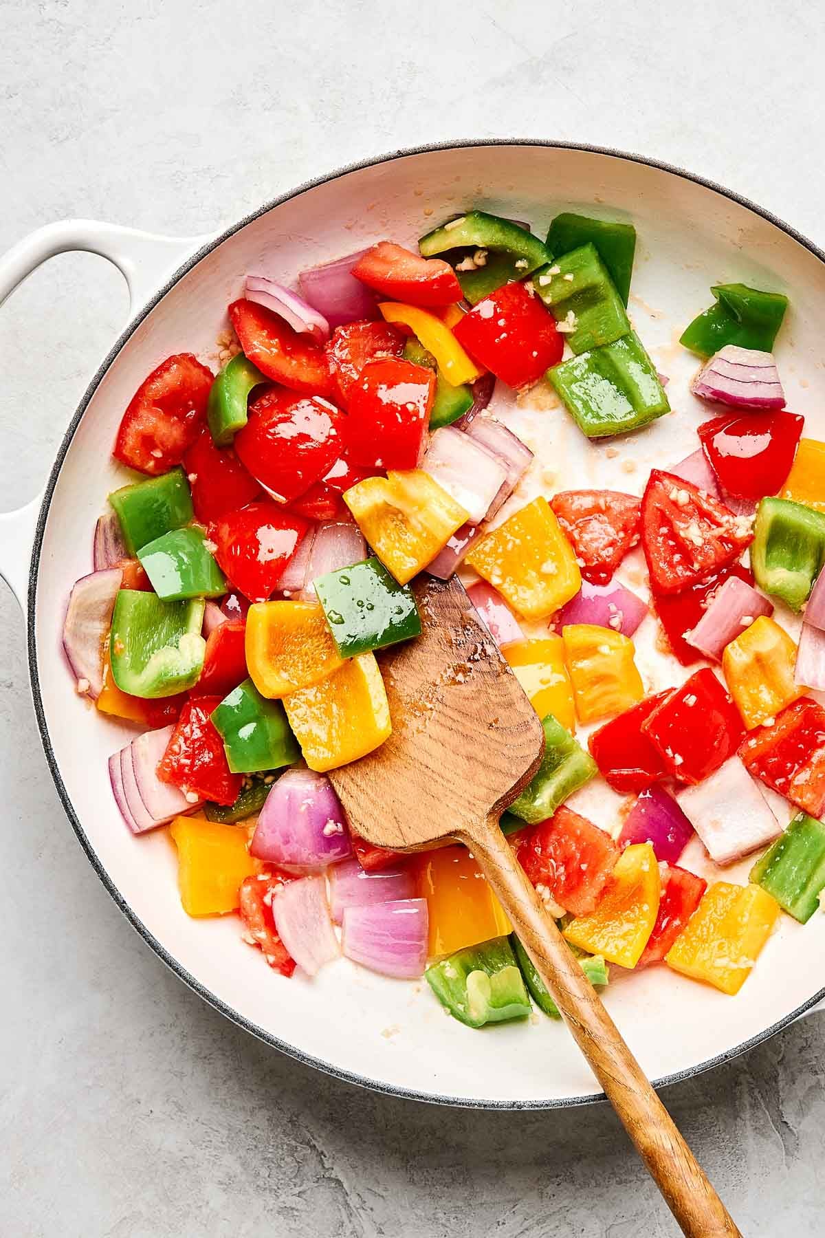 A white skillet with chopped red, green, and yellow bell peppers, red onions, and tomatoes being sautéed with a wooden spatula on a light surface.