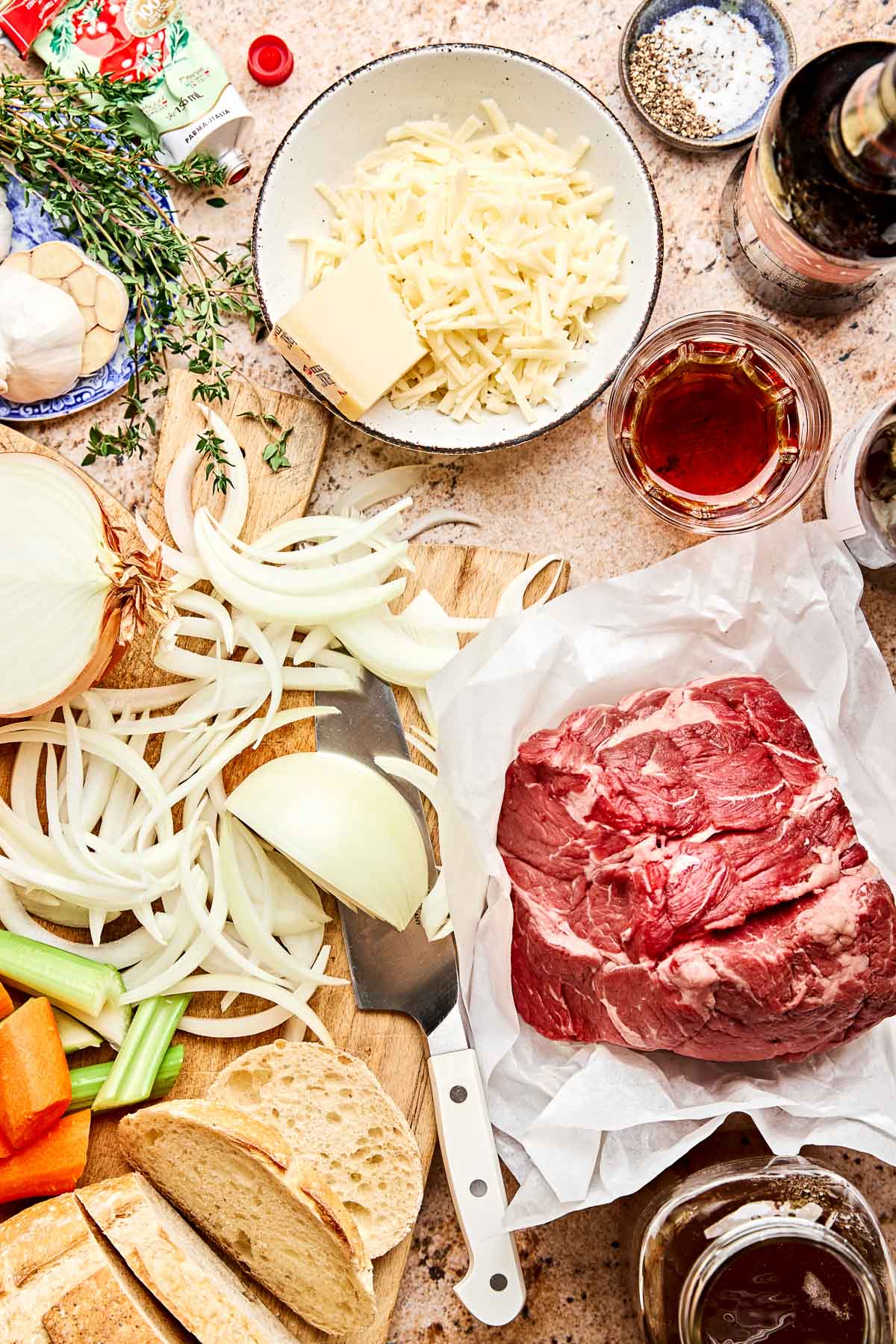 An overhead view of a kitchen counter with ingredients displayed: raw beef, sliced onions, celery, carrots, grated cheese, butter, fresh herbs, bread, garlic, seasonings, and bottles of wine and broth.