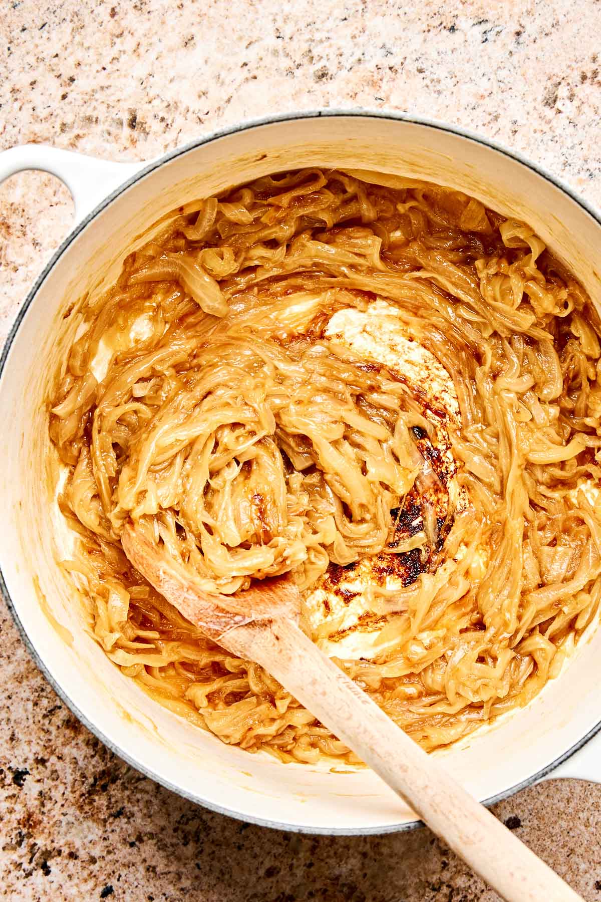A pot filled with caramelized onions being stirred with a wooden spoon on a speckled countertop. The onions are golden brown and creamy, with some browning on the bottom of the pot.