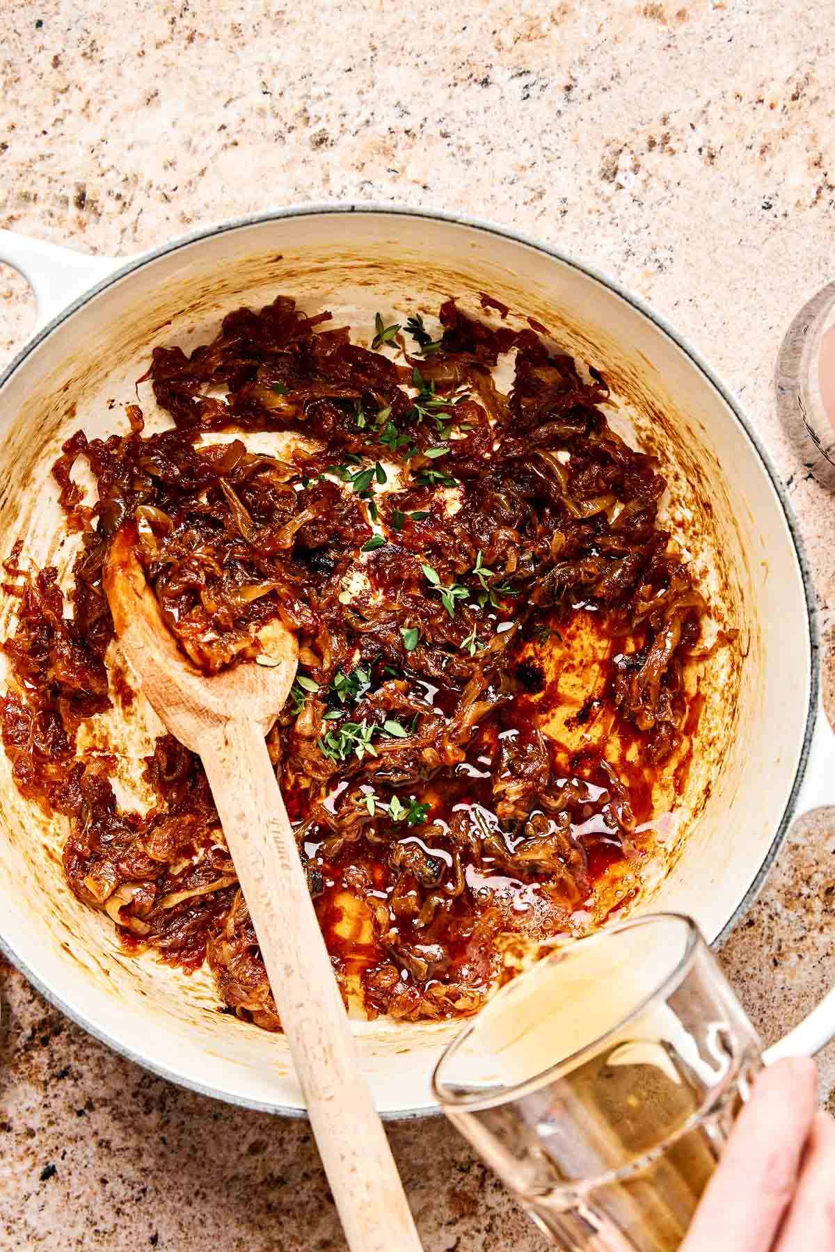 A white pot with caramelized onions, fresh thyme, and a wooden spoon on a speckled countertop. A hand pours liquid from a glass into the pot.