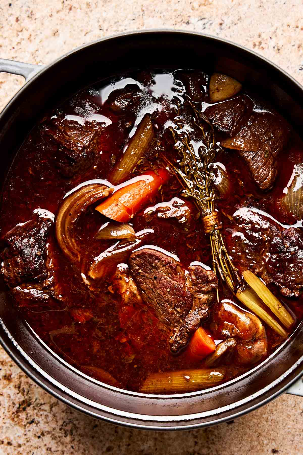 A pot filled with rich, dark French onion soup featuring chunks of beef, carrots, celery, onions, and a bundle of fresh herbs in a savory broth.