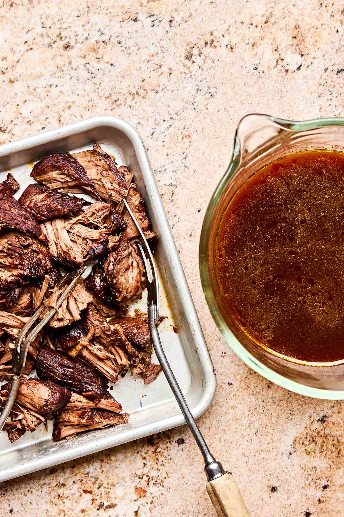 Shredded cooked beef on a metal tray with a meat fork, next to a glass measuring cup filled with brown beef broth, all on a light stone countertop.