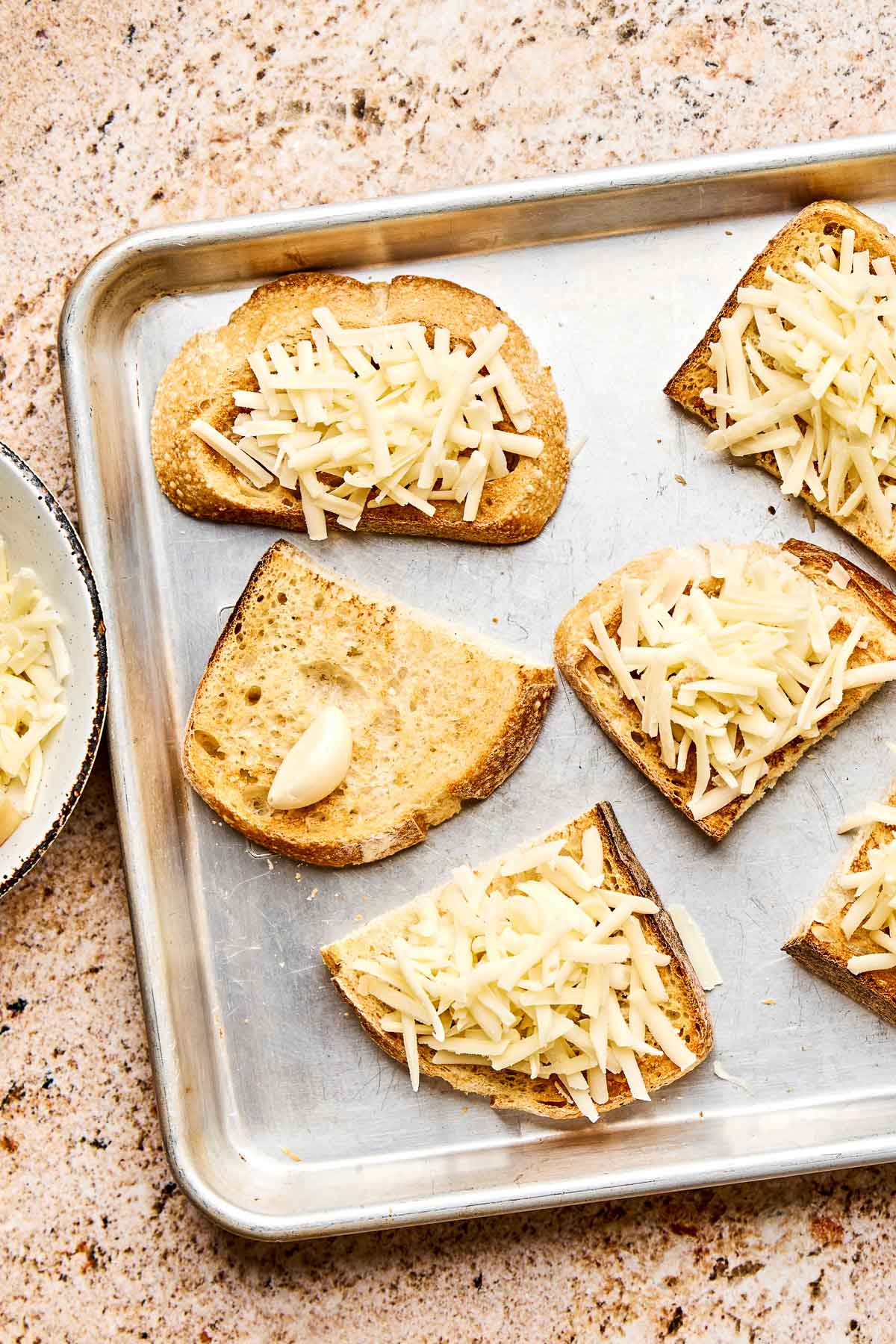 A baking tray with six slices of bread, five topped with shredded cheese and one with a garlic clove. The tray sits on a speckled countertop, and a bowl of shredded cheese is partially visible to the side.