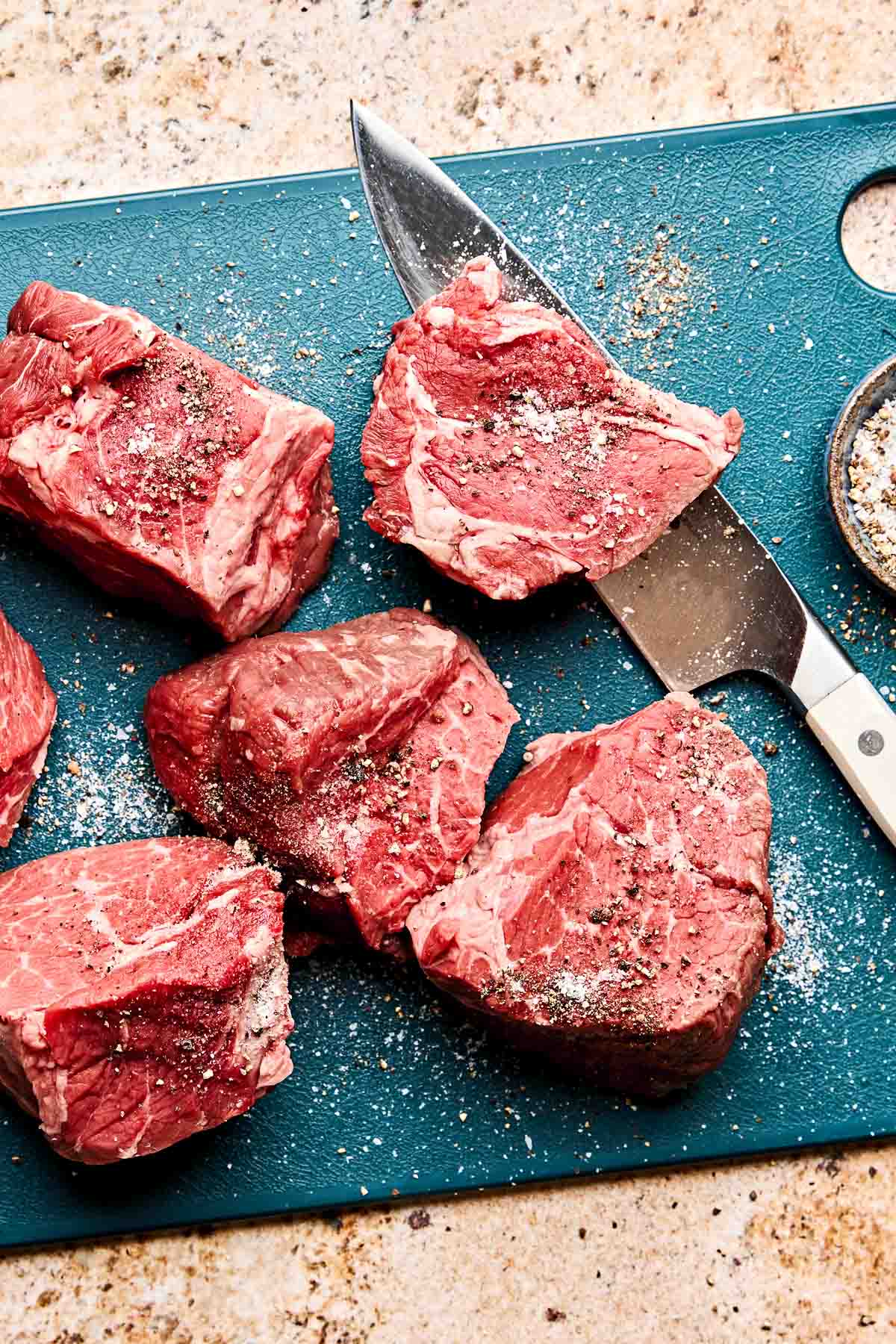 Raw filet mignon steaks seasoned with salt and pepper sit on a blue cutting board next to a knife and a small bowl of seasoning on a speckled countertop.