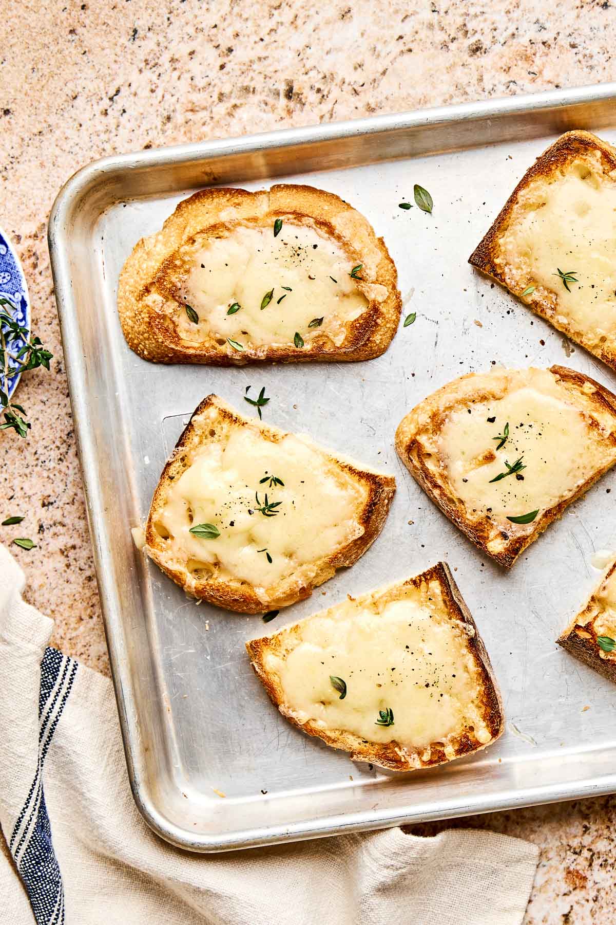 Six slices of toasted bread topped with melted cheese and garnished with fresh herbs are arranged on a metal baking sheet, resting on a light-colored countertop.