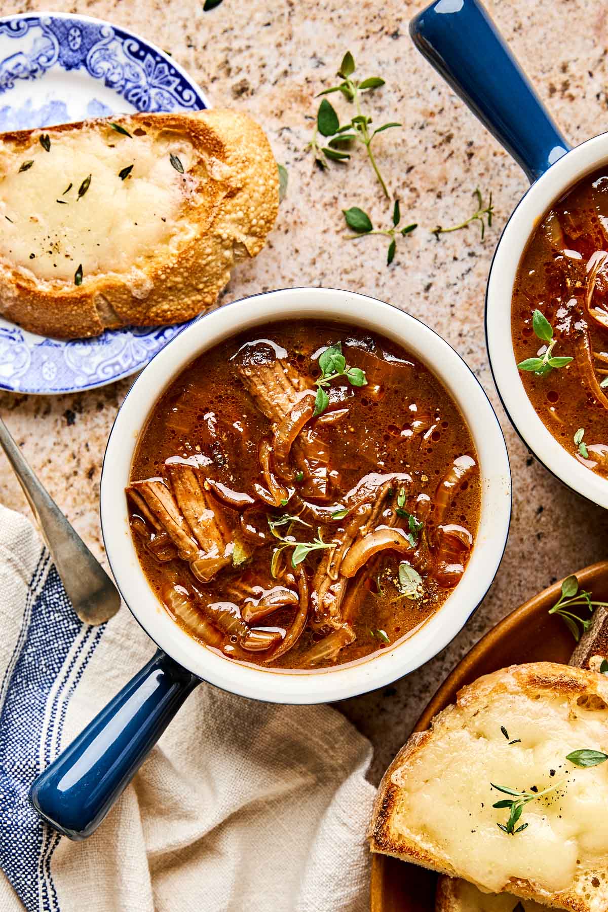 Two bowls of French onion soup with braised beef, topped with fresh herbs. Nearby is a slice of toasted bread with melted cheese on a blue plate, sitting on a beige countertop with scattered thyme leaves.