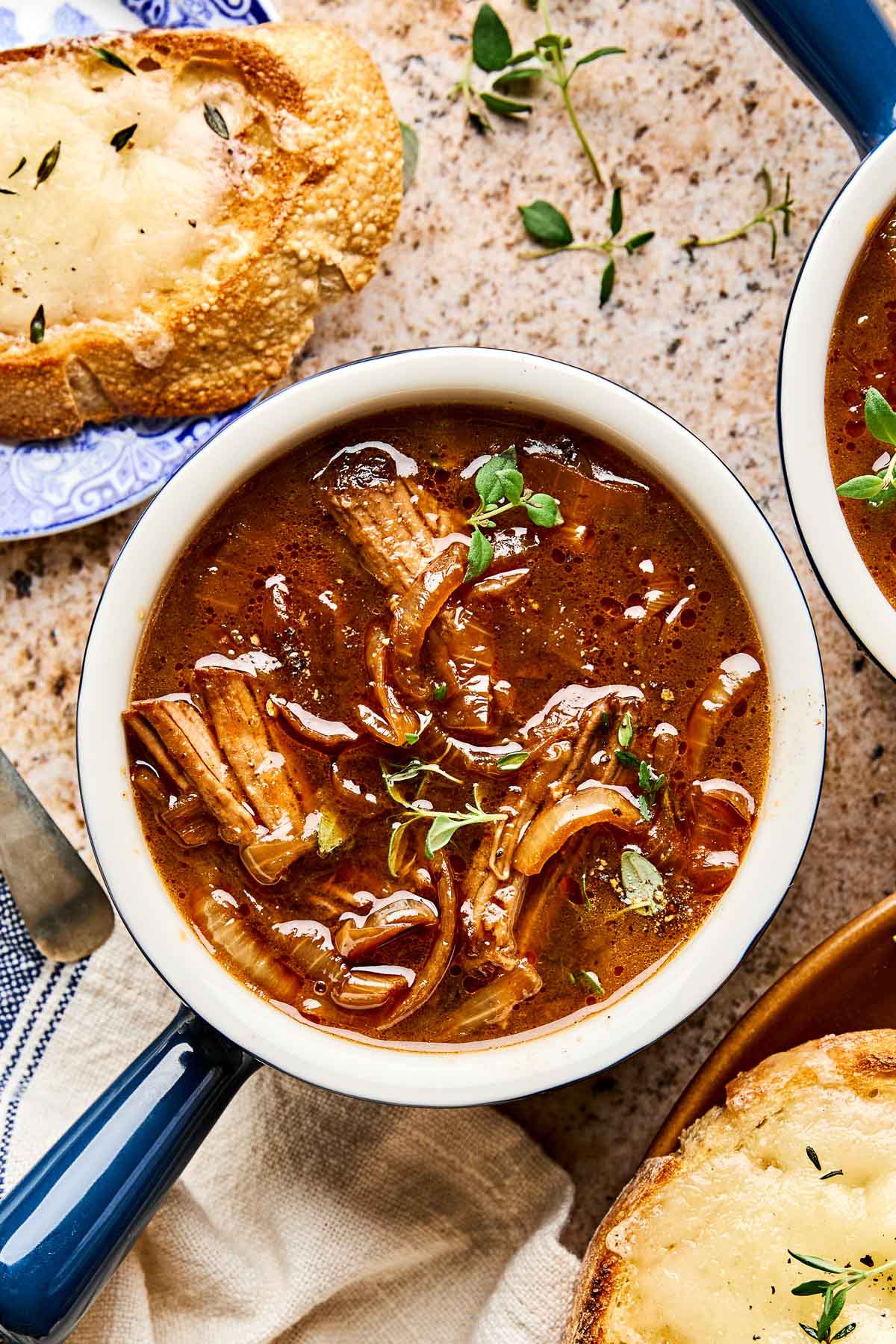 A bowl of beef French onion soup topped with fresh herbs, surrounded by slices of toasted bread with melted cheese on a light countertop.