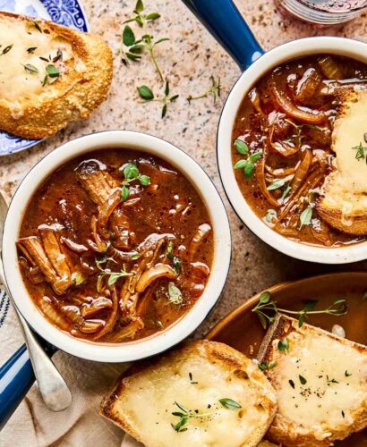 Two bowls of beef French onion soup topped with fresh herbs, served with toasted bread slices covered in melted cheese, on a stone surface with a glass of red wine and a blue-patterned plate.