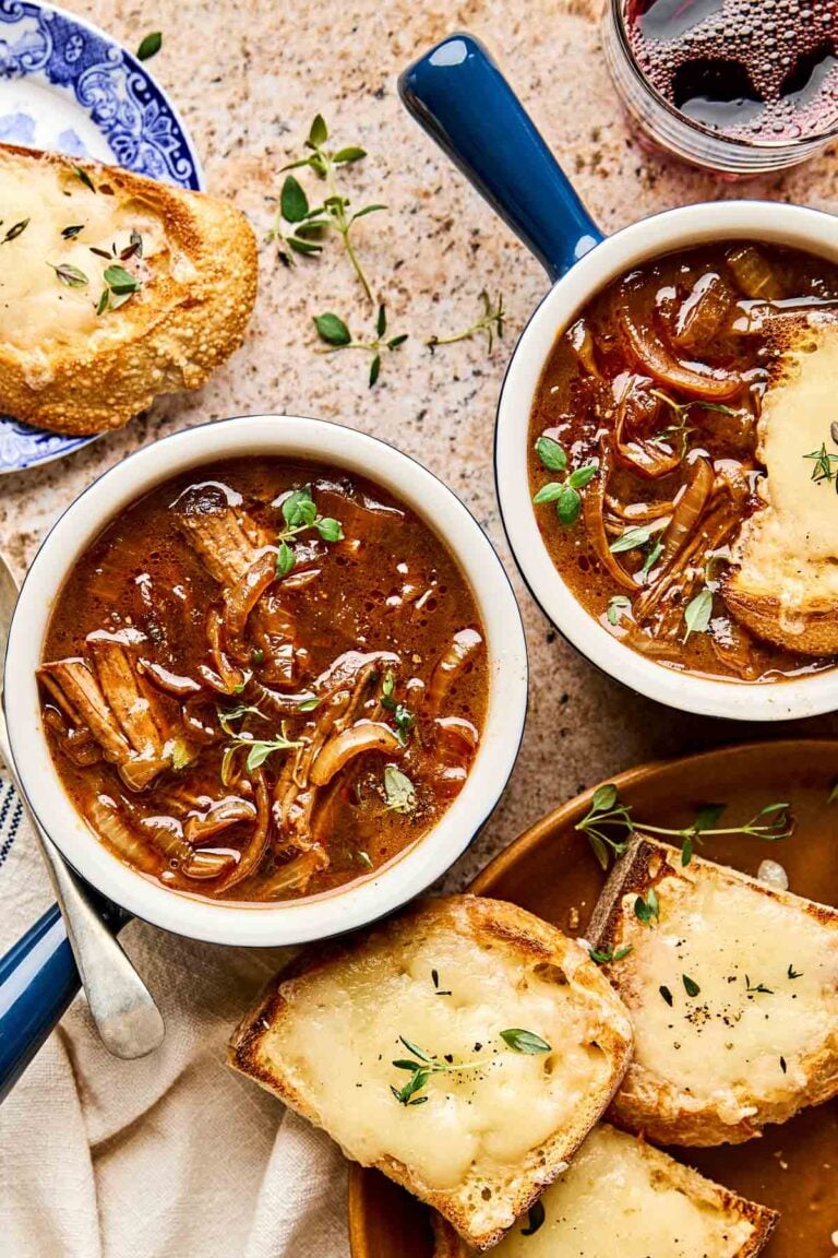 Two bowls of beef French onion soup topped with fresh herbs, served with toasted bread slices covered in melted cheese, on a stone surface with a glass of red wine and a blue-patterned plate.