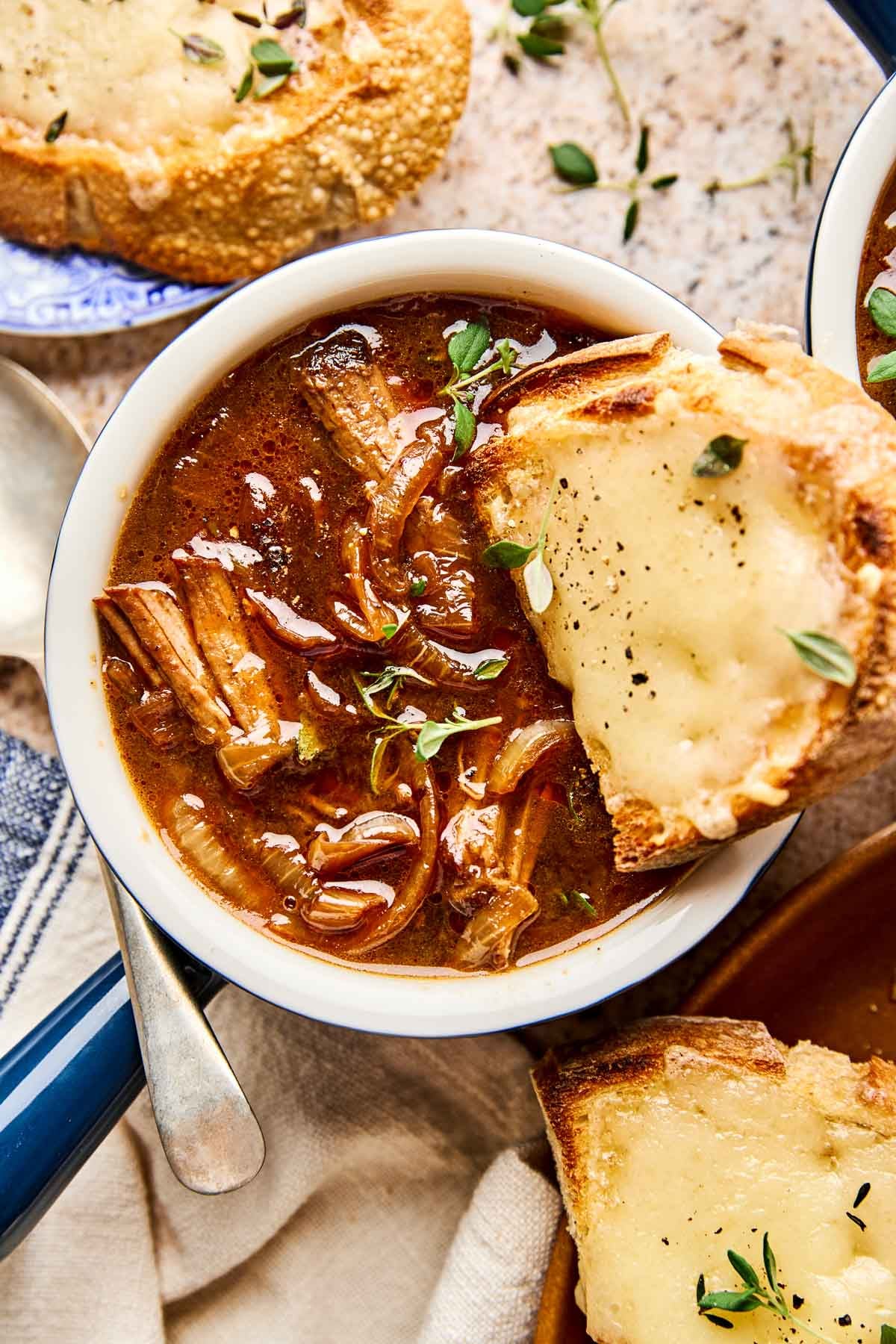 A bowl of beef French onion soup with caramelized onions and herbs, served with a slice of toasted bread covered in melted cheese. Another cheesy bread slice and a spoon are nearby.