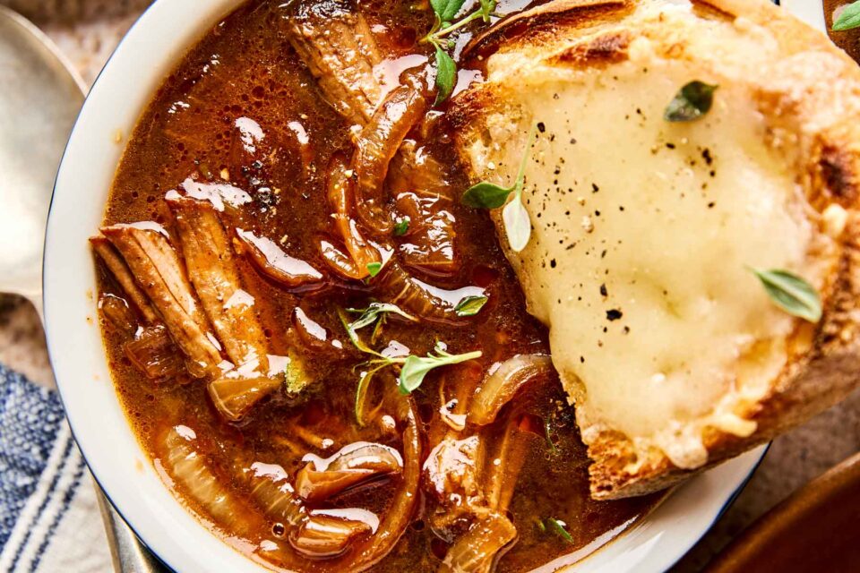 A bowl of French onion soup with caramelized onions and shredded beef in rich broth, garnished with fresh herbs, served with a slice of crusty bread topped with melted cheese and black pepper.