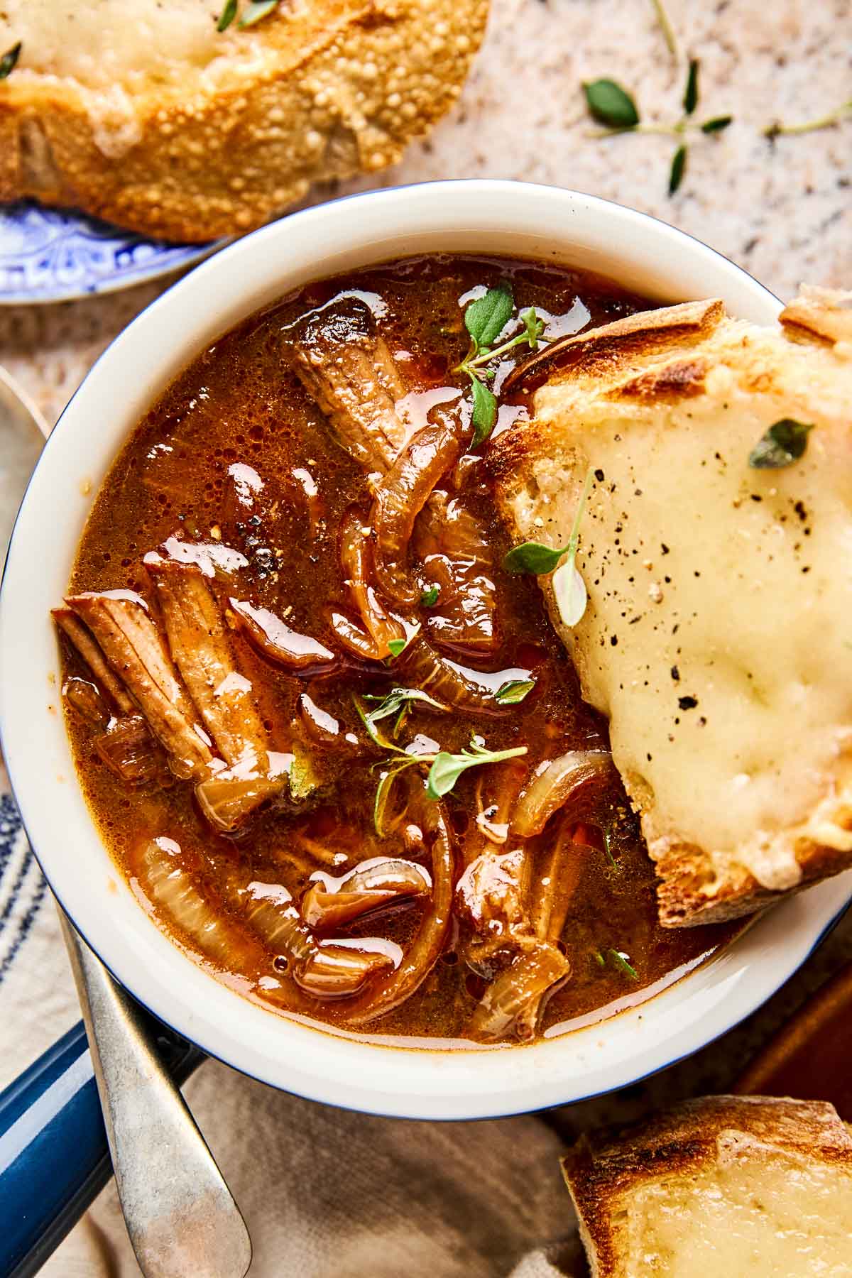 A bowl of French onion soup with caramelized onions and beef, topped with fresh herbs, served with a slice of toasted bread covered in melted cheese. Another cheese toast is visible in the background.