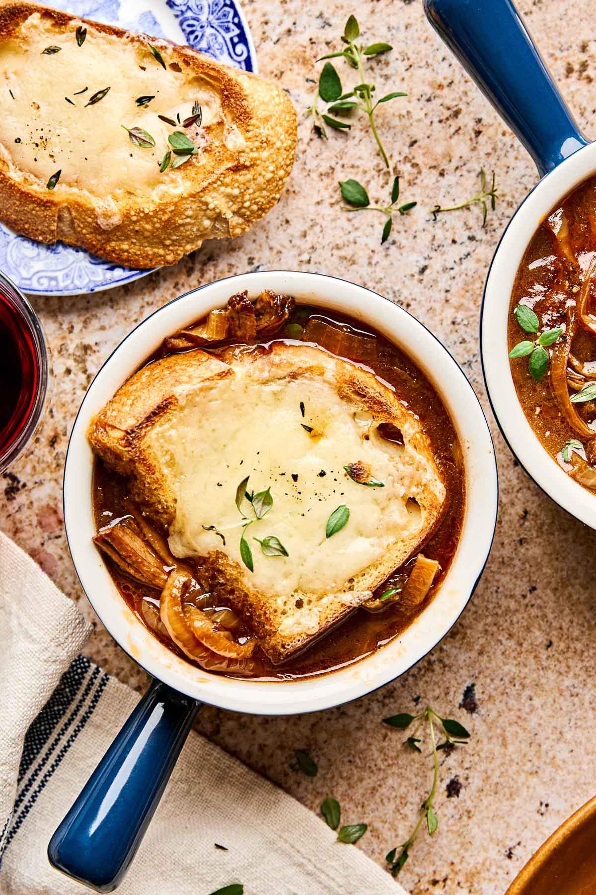 A bowl of beef French onion soup topped with a slice of toasted bread and melted cheese, garnished with fresh thyme. Another bowl and a slice of bread are partially visible nearby on a stone countertop.