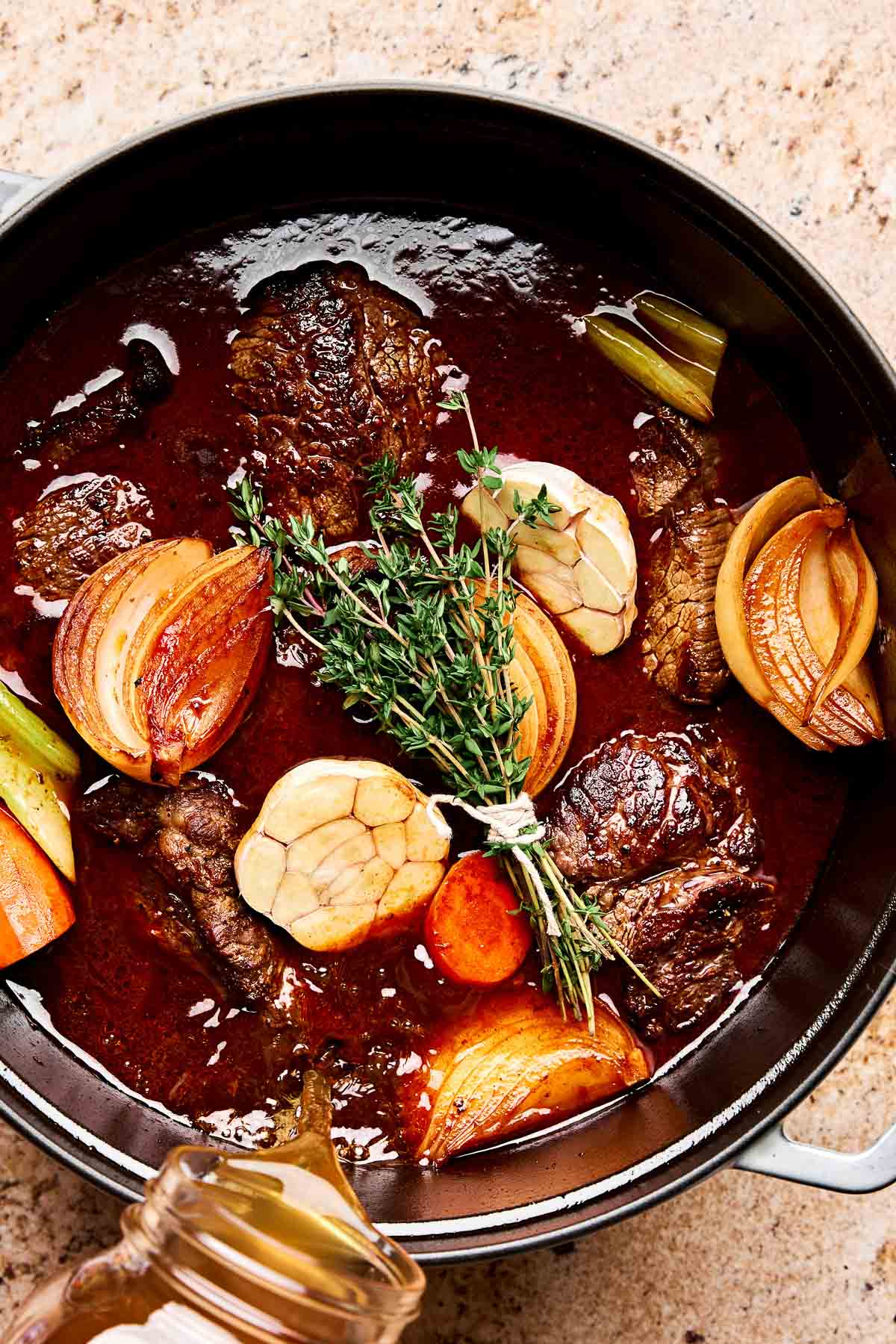 A pot of braised beef with rich brown sauce, topped with halved garlic bulbs, onions, carrots, celery, and a bundle of fresh thyme, on a beige countertop.