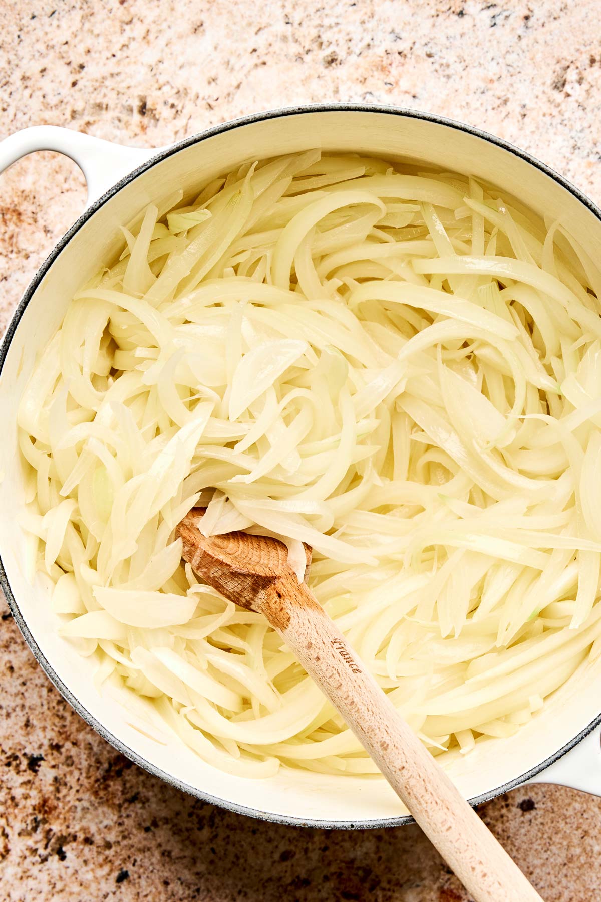 A white pot filled with sliced raw onions being stirred with a wooden spoon, set on a speckled countertop.
