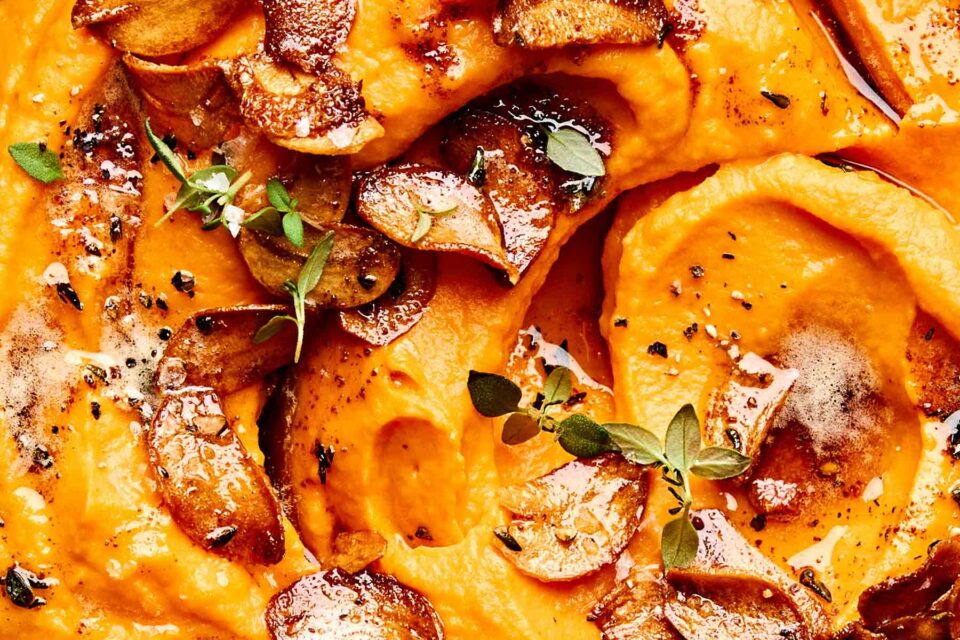 Close-up of creamy mashed sweet potatoes topped with browned butter, crispy garlic slices, fresh thyme leaves, and black pepper. The vibrant orange mash contrasts with the golden garlic and green herbs.