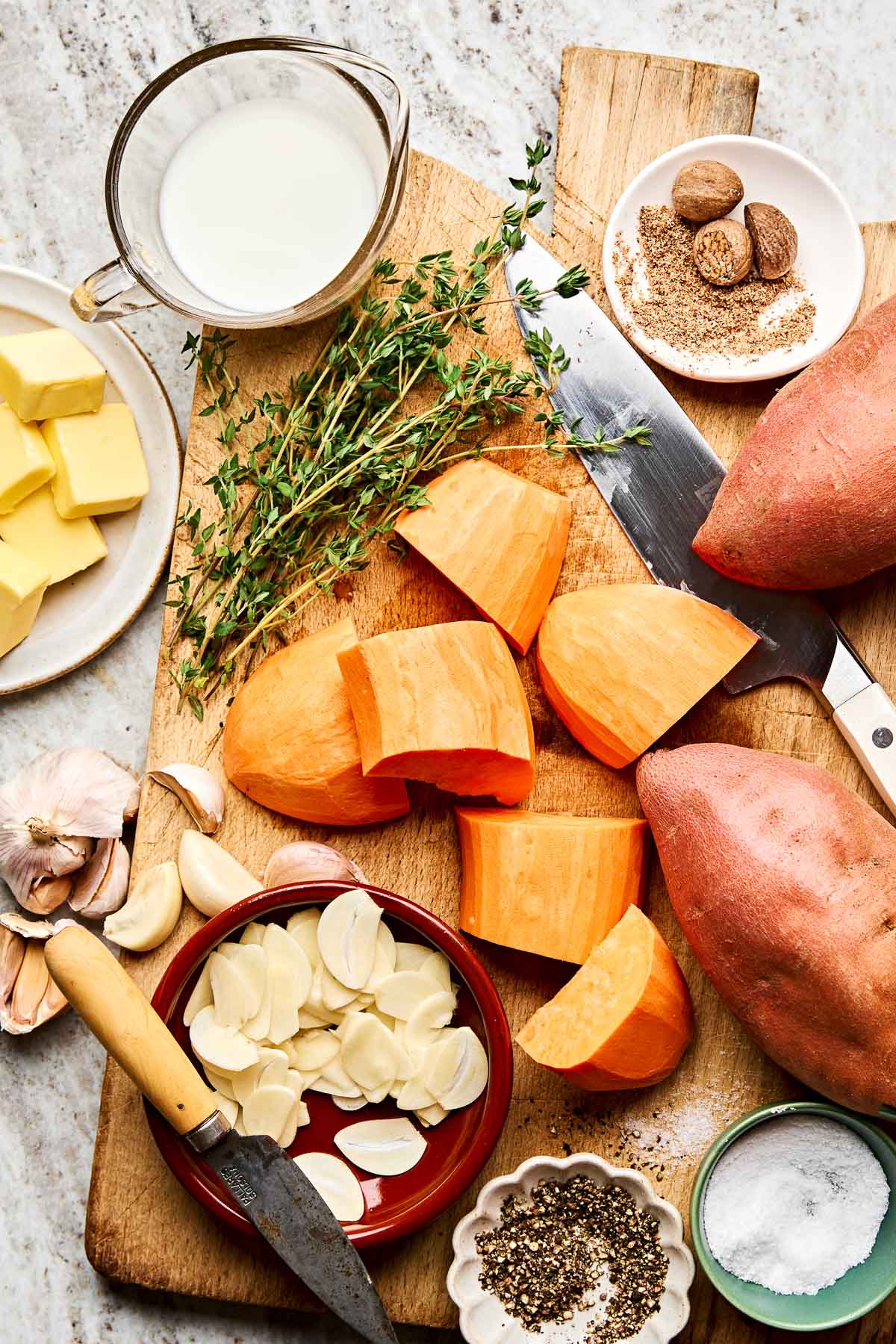 Chopped sweet potatoes, fresh thyme, sliced garlic, butter, ground spices, milk, salt, and pepper on a wooden cutting board with a knife, ready for cooking.