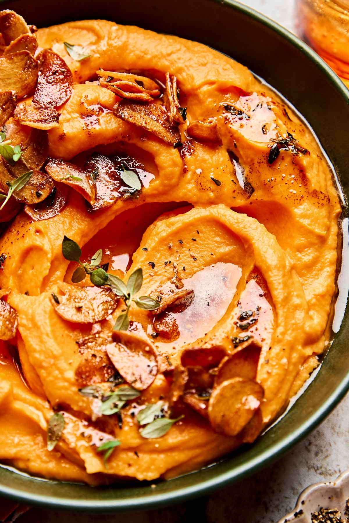 A bowl of creamy whipped sweet potatoes topped with crispy garlic slices, fresh herbs, and a drizzle of oil or butter. The dish is garnished with black pepper and served in a green bowl.
