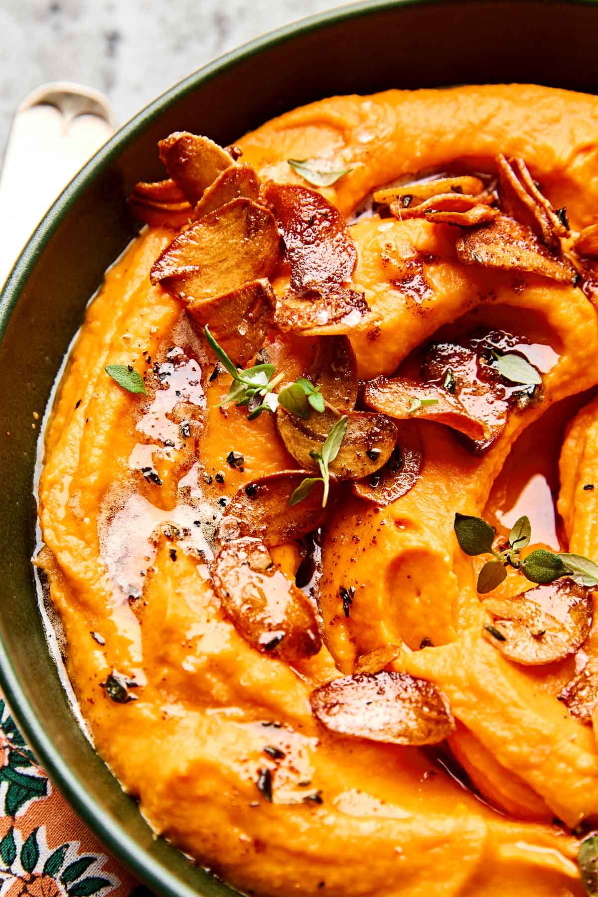 A close-up of creamy whipped sweet potatoes topped with crispy garlic slices, fresh herbs, melted butter, and cracked black pepper in a dark bowl.