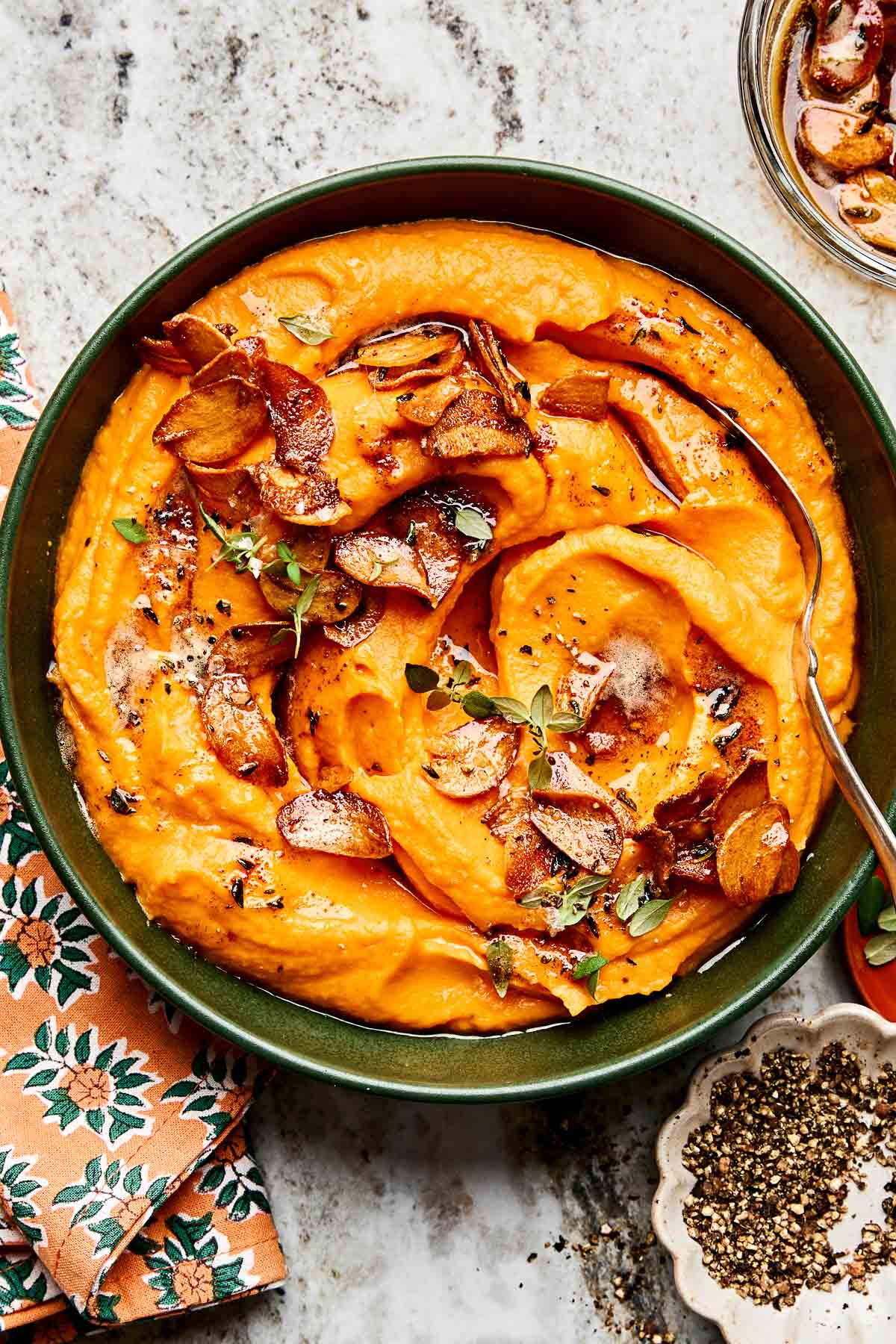 A bowl of creamy mashed sweet potatoes topped with crispy garlic slices and fresh herbs, with a spoon resting in the bowl. A floral napkin and a small dish of ground black pepper sit nearby.