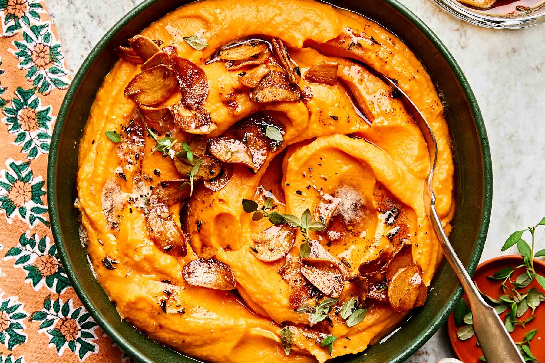 A bowl of creamy sweet potato mash topped with crispy garlic slices, fresh herbs, and black pepper, with a spoon resting inside. A floral napkin and a small bowl are nearby.