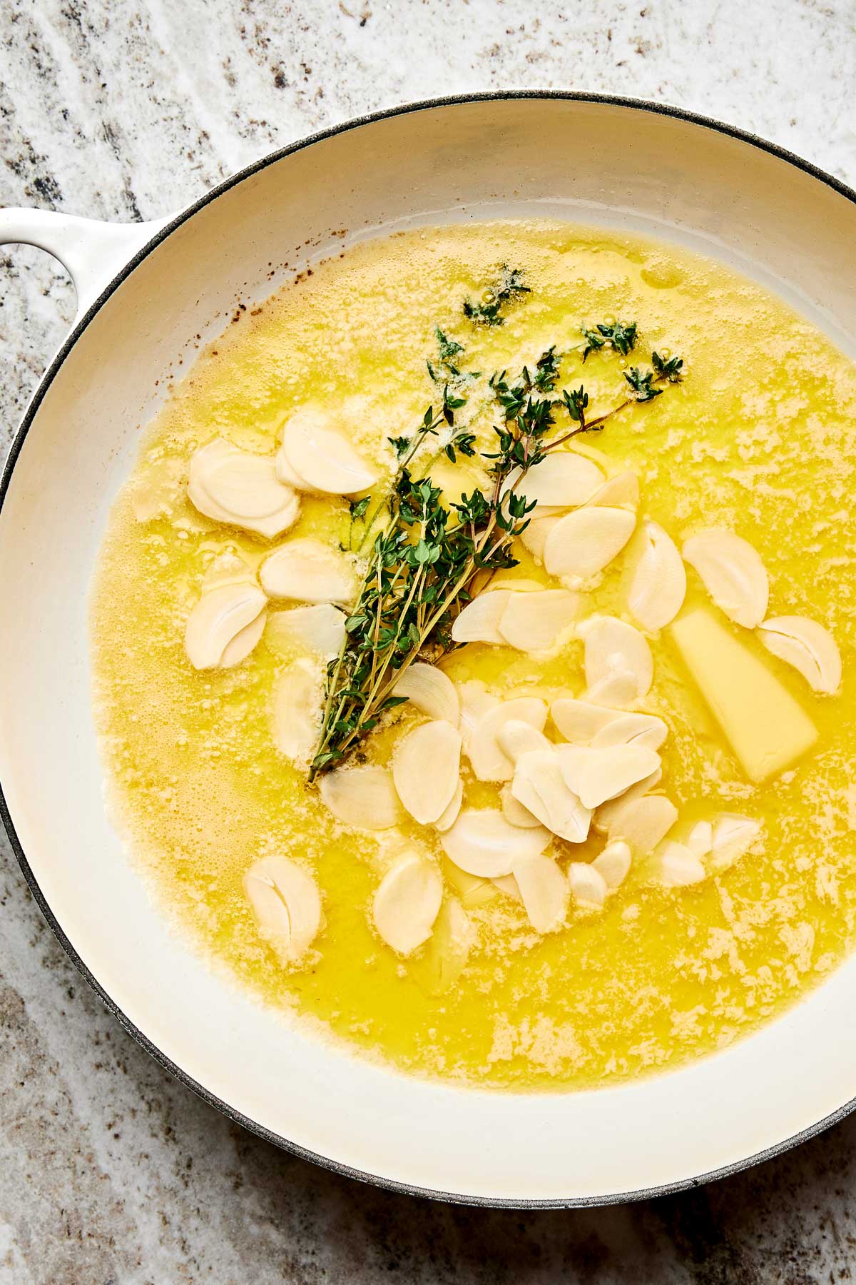 A white pan with melted butter, sliced garlic, and fresh thyme sprigs on a light countertop.