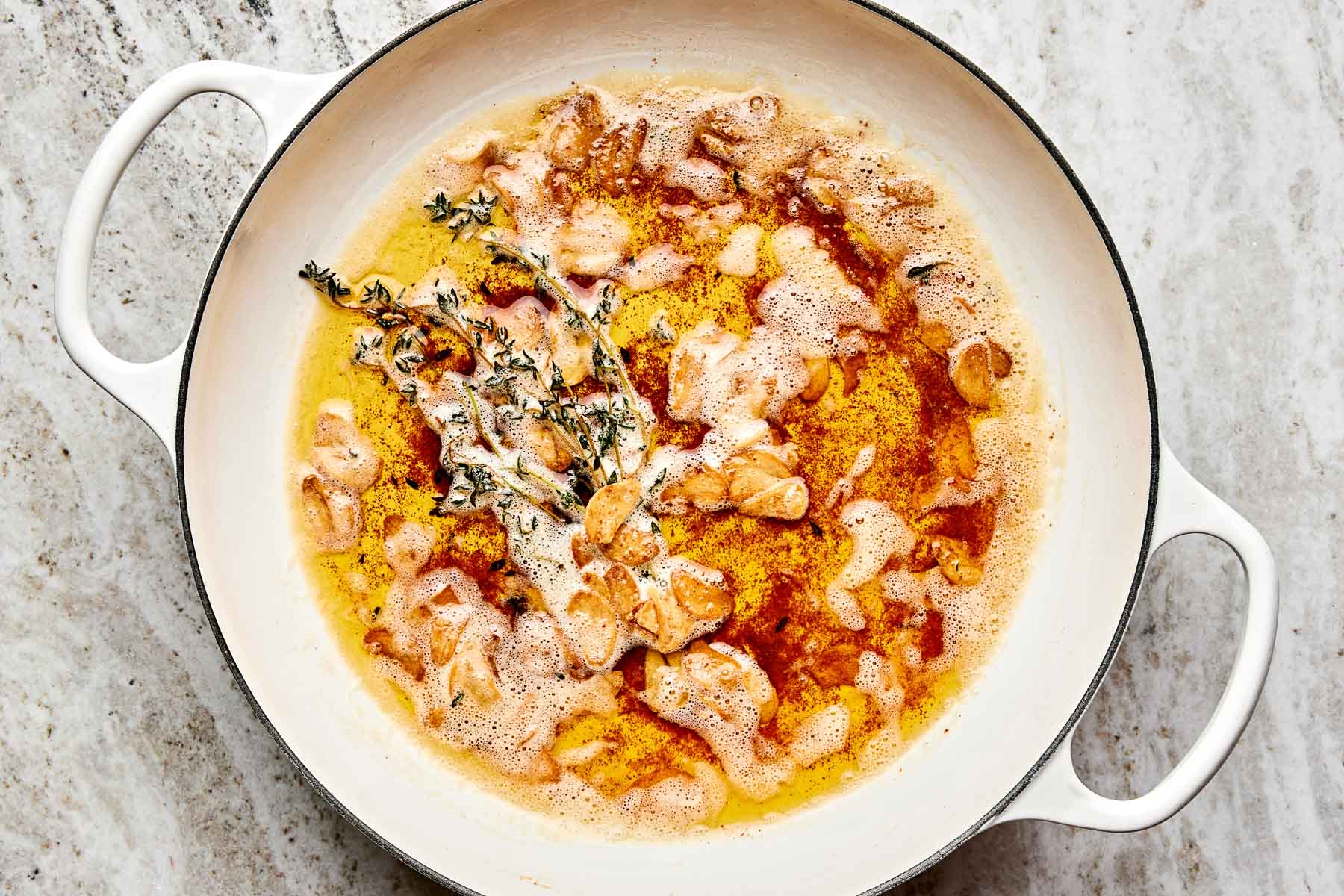 A white enamel pot on a marble counter contains sizzling bacon pieces in bubbling fat, with sprigs of fresh thyme on top.