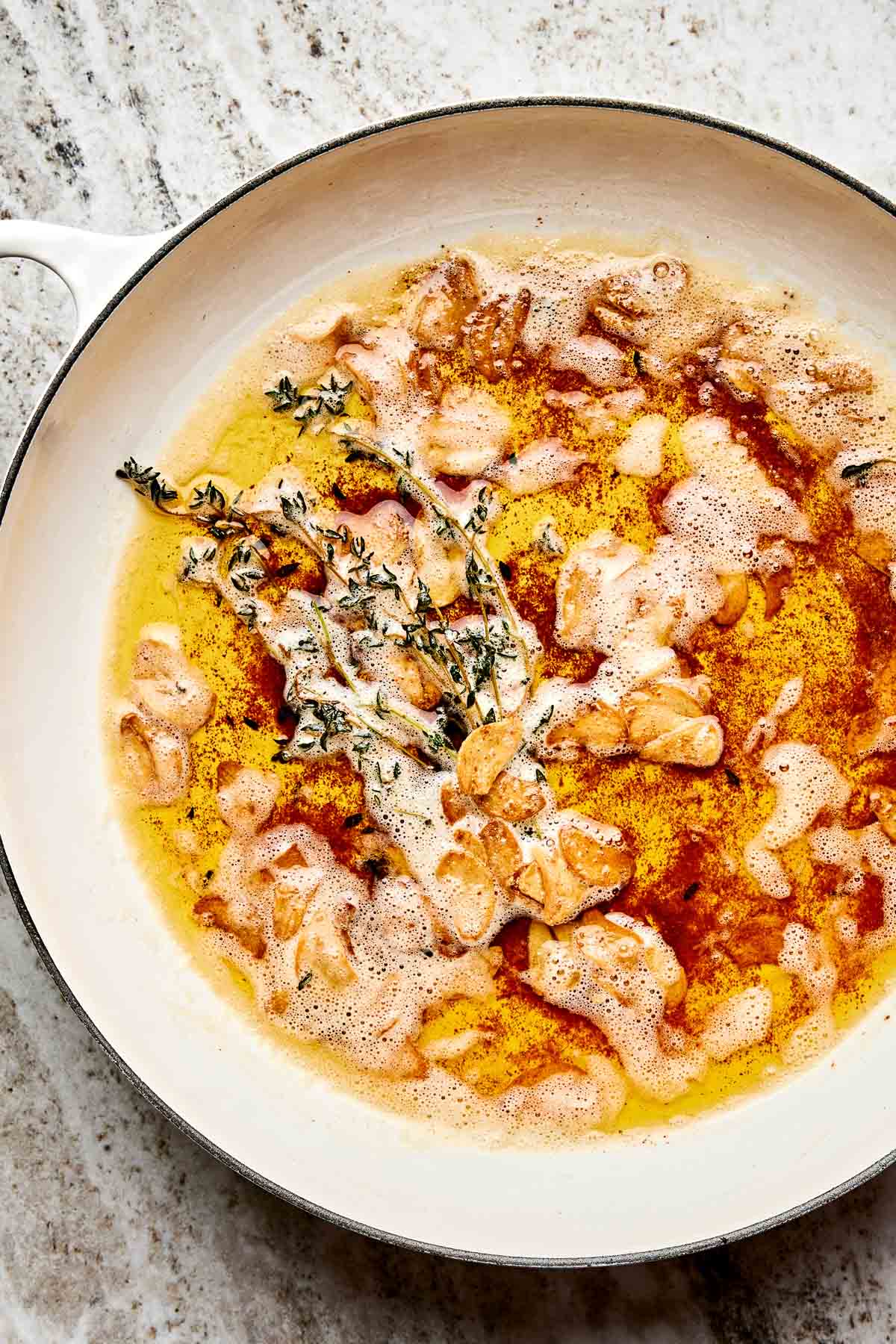 A white skillet on a light countertop filled with browned butter, golden garlic slices, and sprigs of fresh thyme.