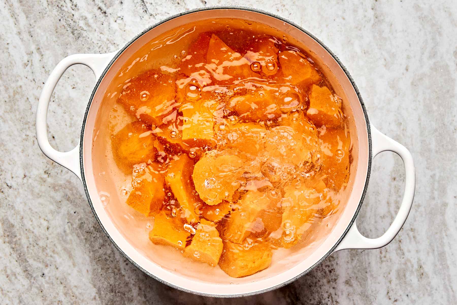 A white pot filled with chunks of orange sweet potatoes boiling in water, placed on a light-colored marble surface.