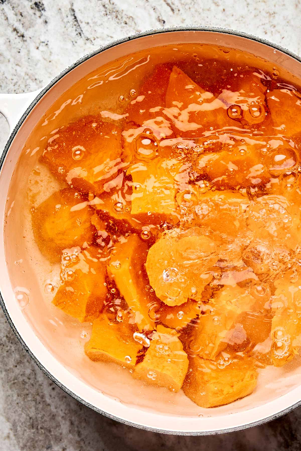 Chunks of sweet potatoes boiling in a pot of water on a light-colored countertop. Bubbles are visible on the surface as the vegetables cook.