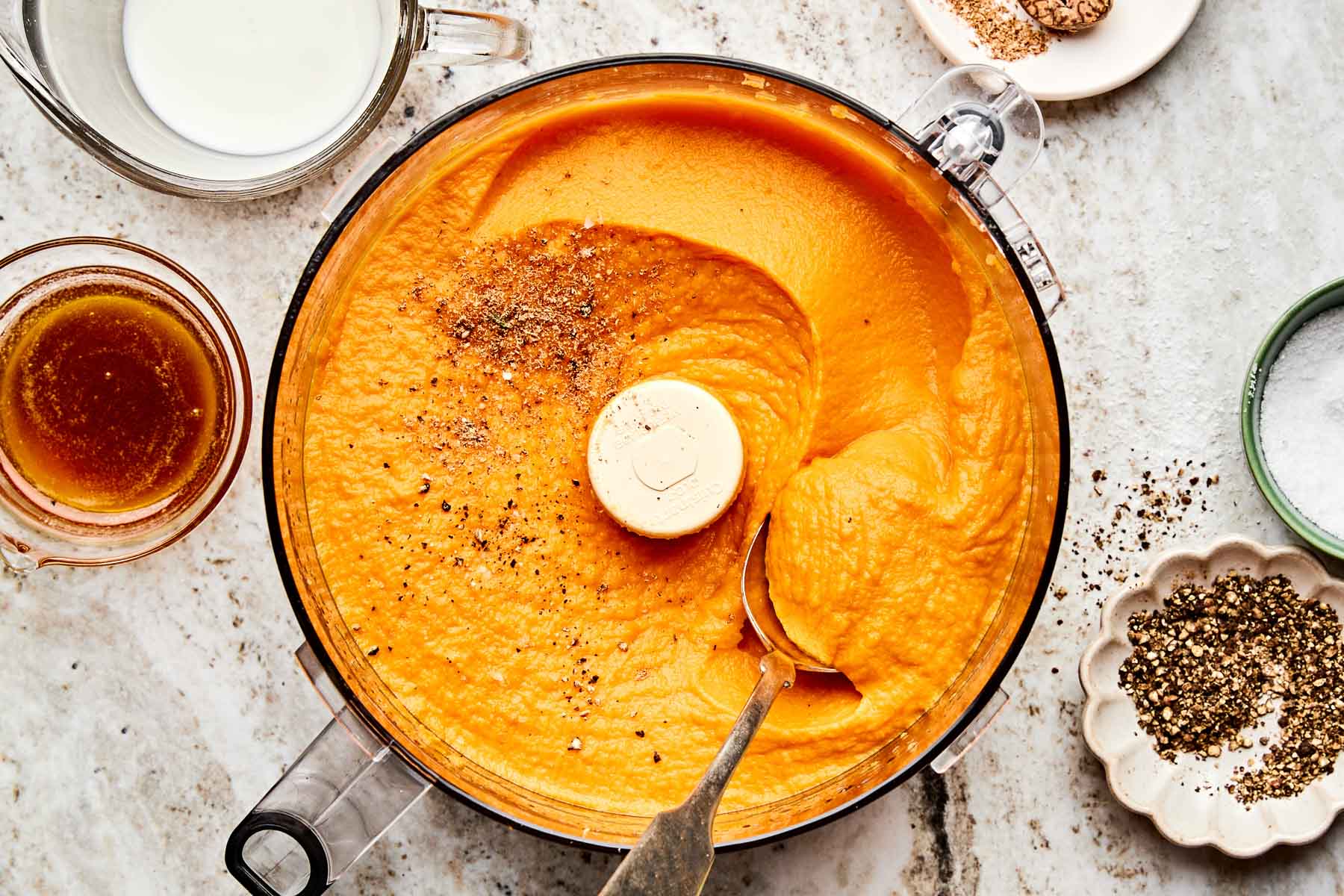 A food processor filled with creamy orange mashed sweet potatoes, topped with cracked black pepper. Surrounding the bowl are ingredients including milk, brown liquid, ground spices, and a dish of black pepper.