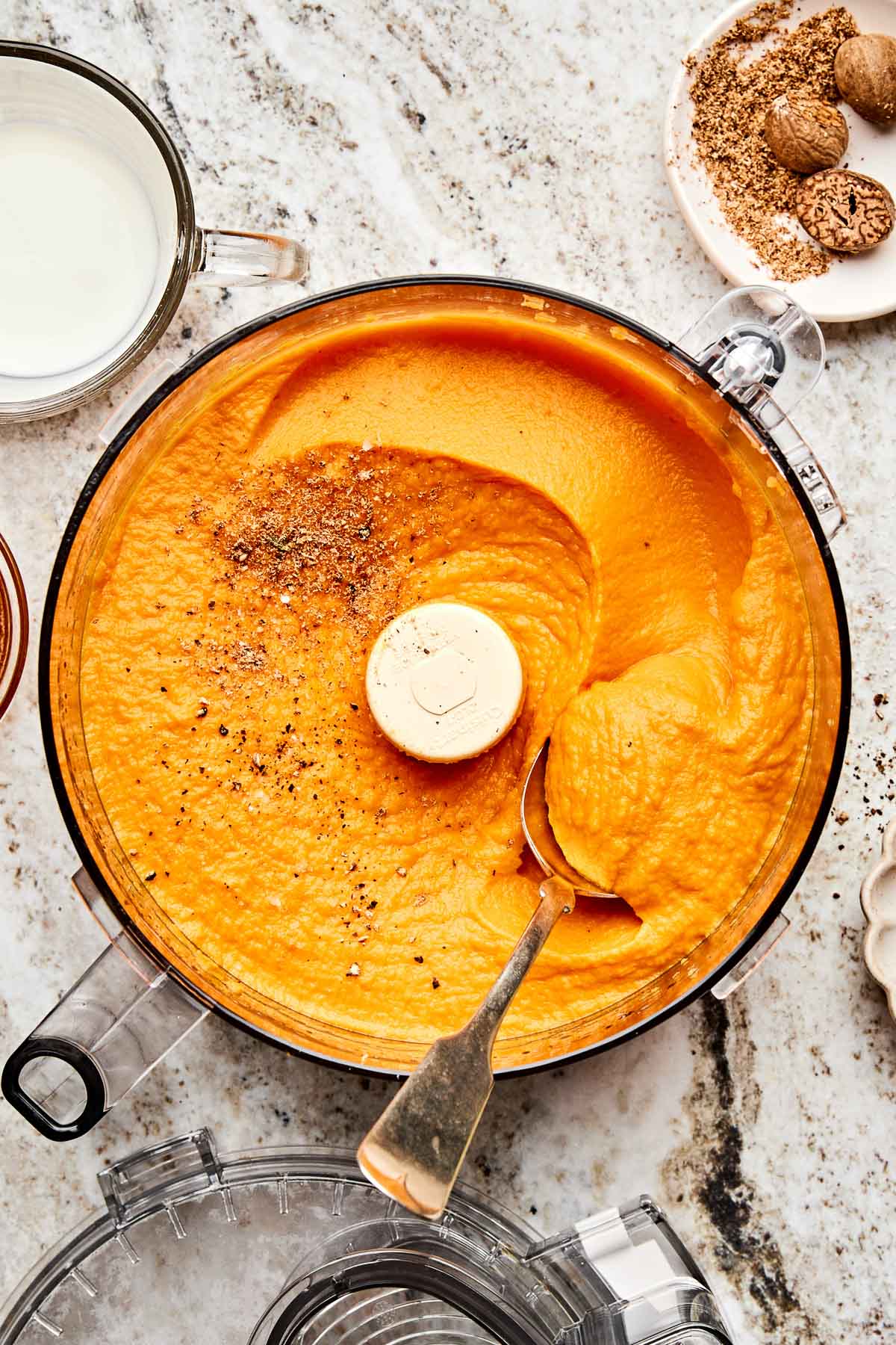 A food processor filled with smooth orange sweet potato puree, topped with ground black pepper. Nearby are a fork, a small bowl of grated nutmeg and whole nutmeg, and a cup of milk on a light marbled countertop.