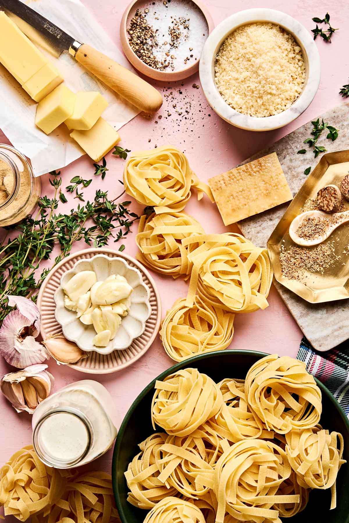 Overhead view of raw fettuccine nests, grated and block parmesan, butter, garlic, heavy cream, figs, black pepper, fresh thyme, and a knife on a pink surface, ready for making a pasta dish.
