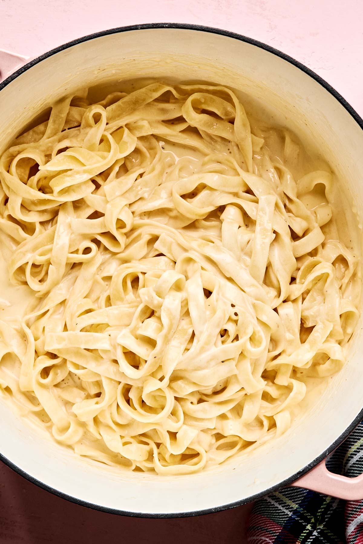 A large pot filled with creamy fettuccine pasta in a white sauce, resting on a light pink surface with part of a checkered cloth visible in the bottom right corner.