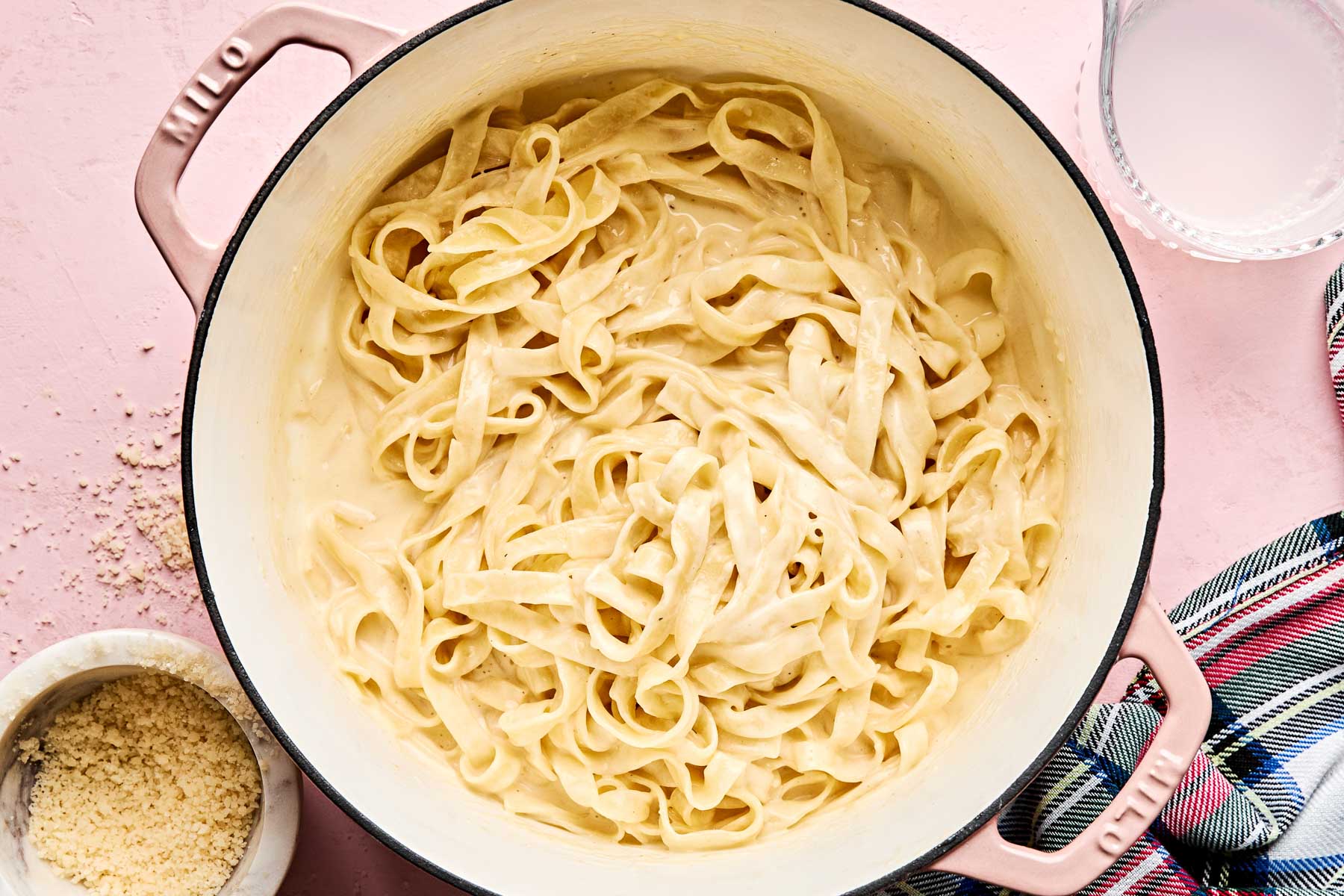 A pot filled with creamy fettuccine sits on a pink surface, next to a bowl of grated cheese, a glass of milk, and a striped kitchen towel.
