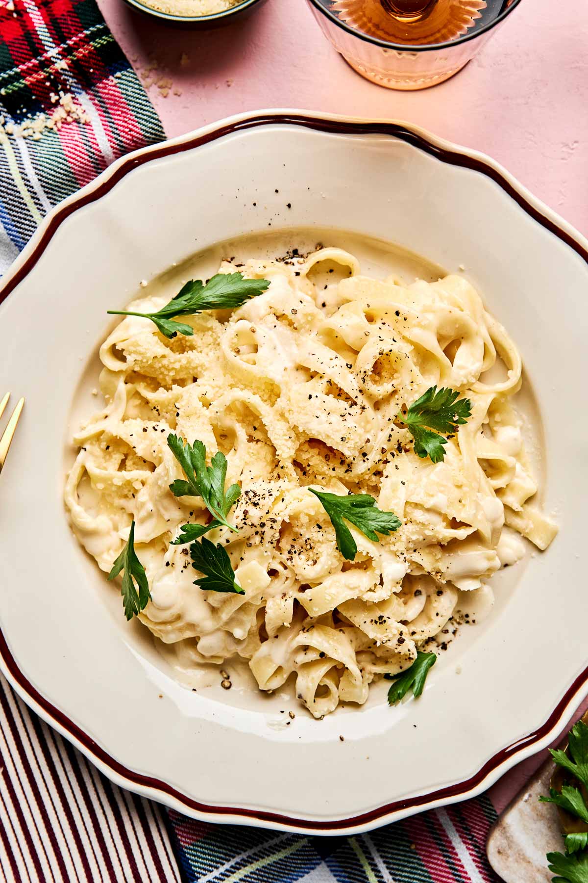 A plate of creamy fettuccine garnished with parsley and cracked black pepper, served on a white dish with a decorative rim, set on a table with a red and green plaid napkin.