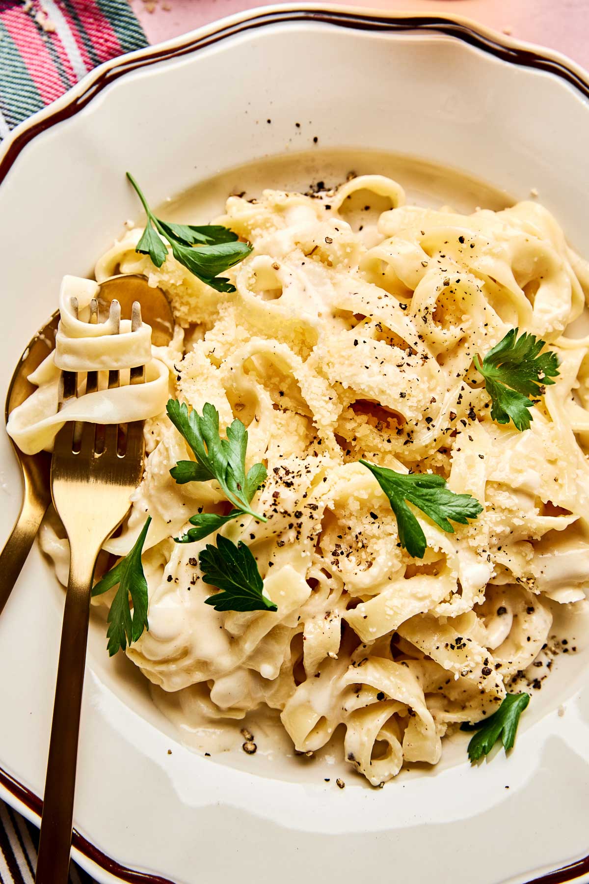 A plate of Christmas fettuccine topped with grated parmesan cheese, black pepper, and fresh parsley, served with a gold fork and spoon on a white plate with a scalloped edge.